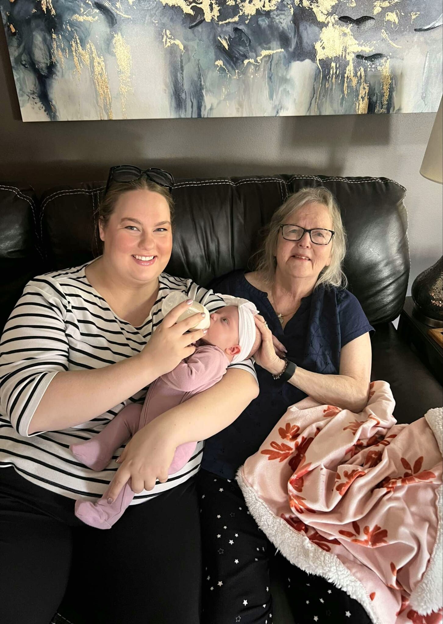 Two women sit on a black leather couch, smiling, as a newborn baby wrapped in pink rests in the younger woman's arms used with a floral blanket nearby—appearing to be a family moment.