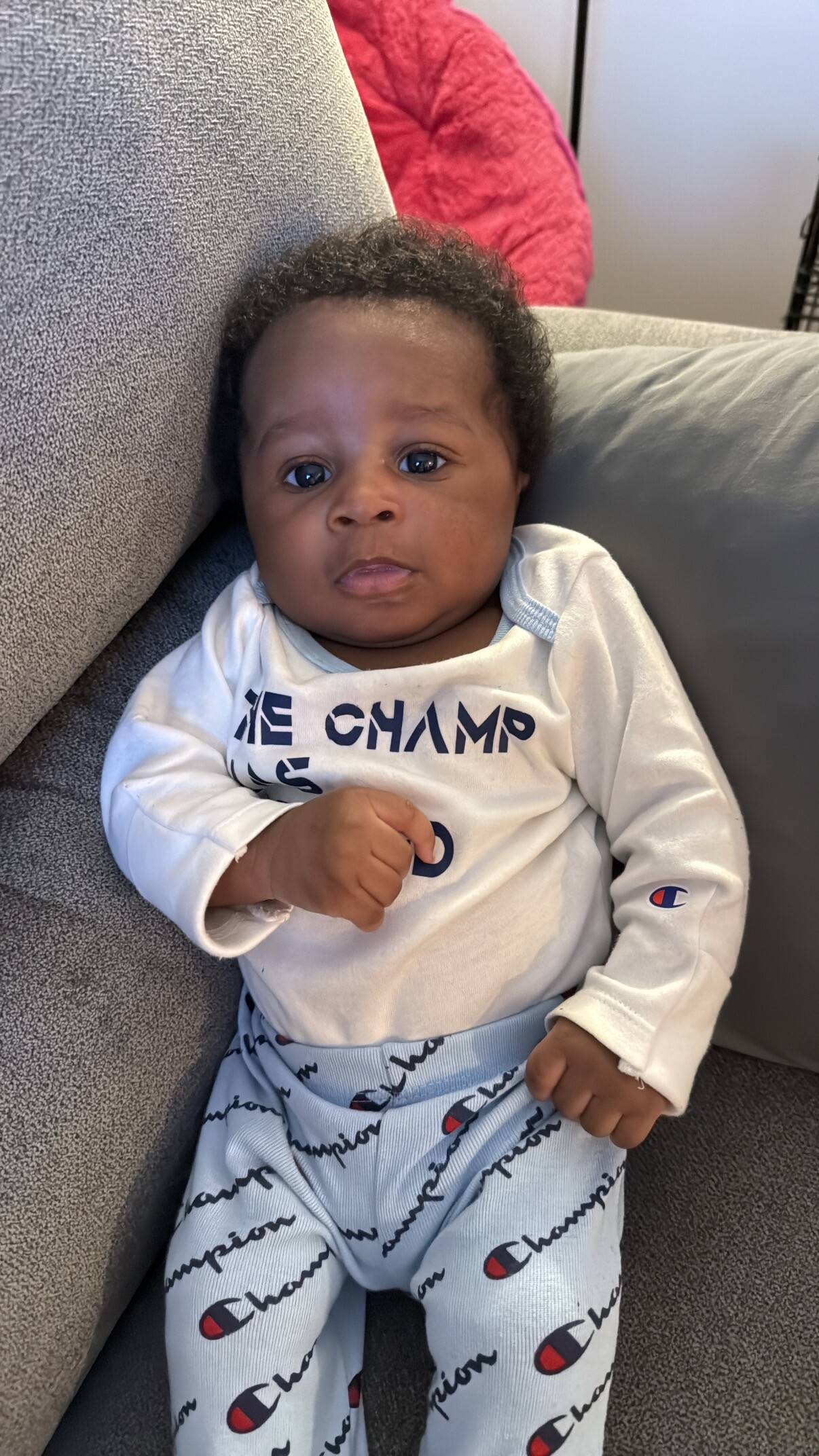 Infant lying on a gray sofa, wearing a white top and light blue Champion pants, looking at the camera with a calm expression.