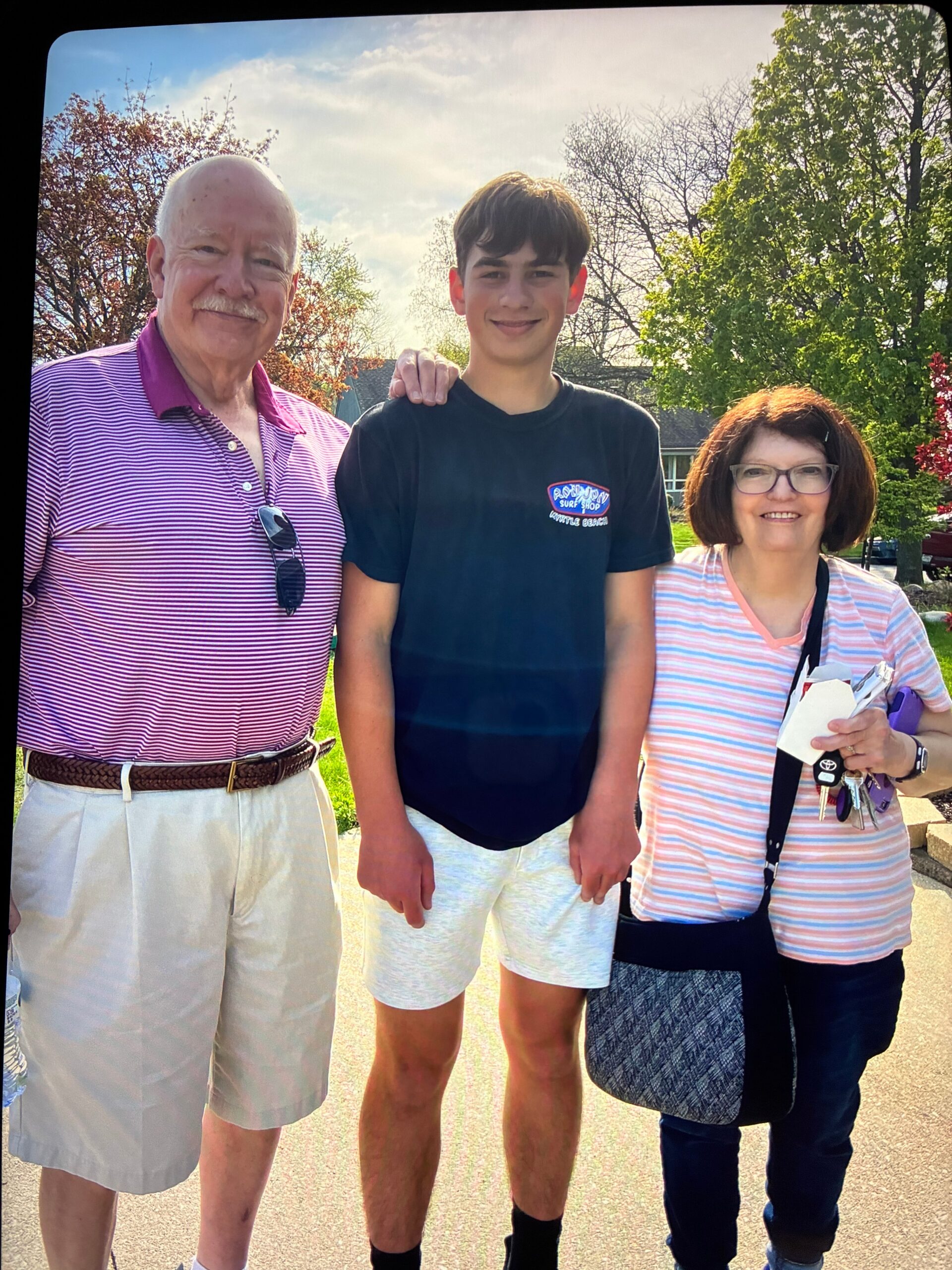 Three people stand together outside, smiling: an older man in a pink striped polo on the left, a teenage boy in a black T-shirt in the middle, and a woman with glasses in a pink striped top on the right, with trees in the sunny background.