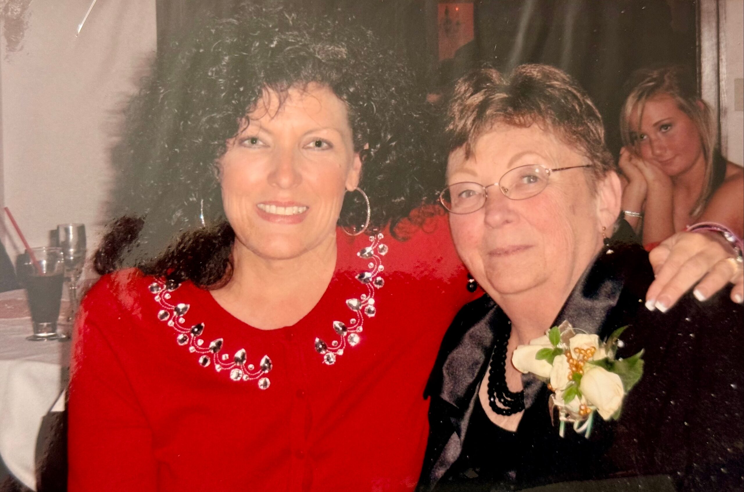 Two women posing at a celebration: one in a red beaded dress, the other wearing glasses with a corsage, and a younger woman in the background.