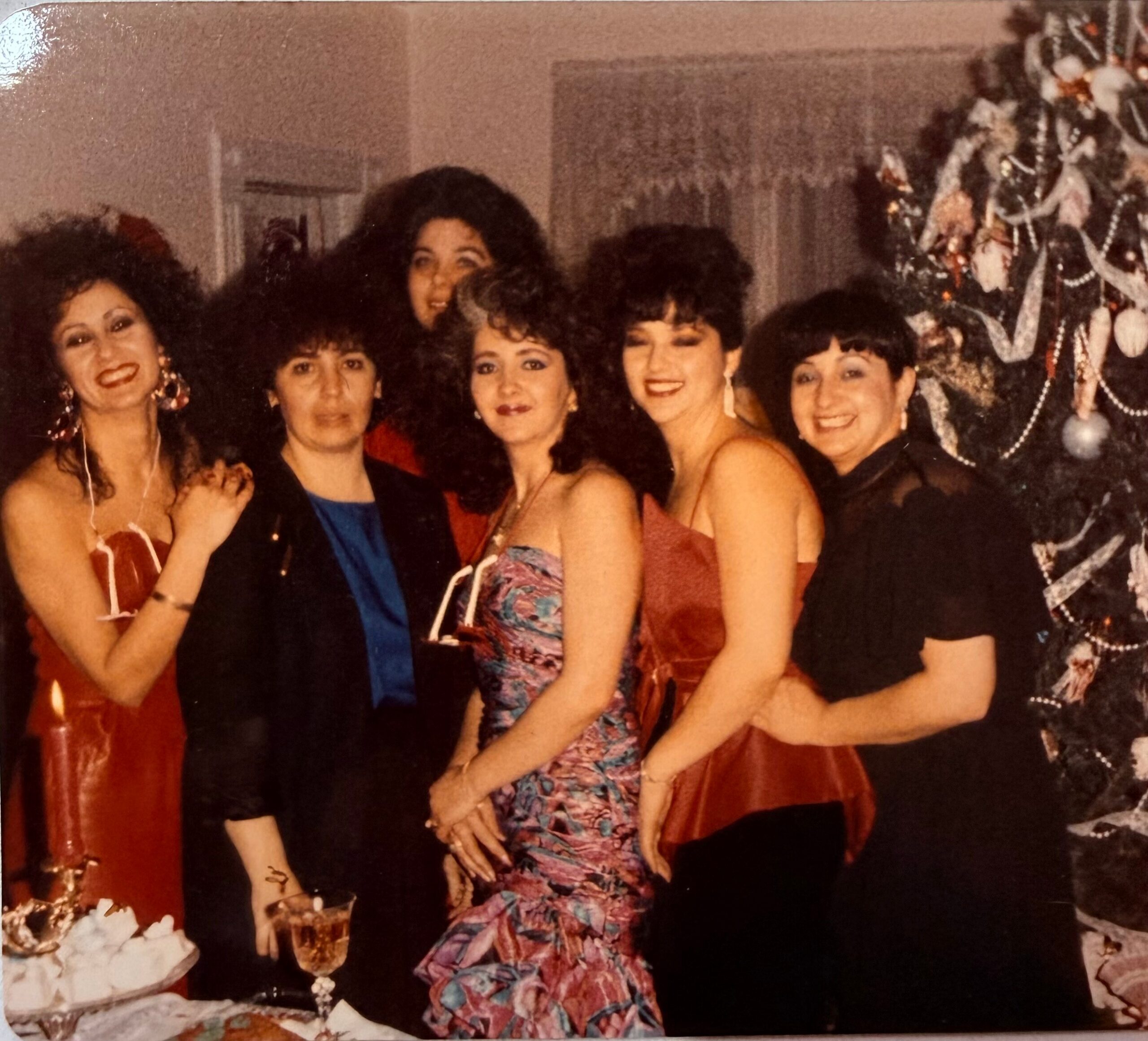 Group of women in formal dresses posing together at a holiday party beside a decorated Christmas tree.