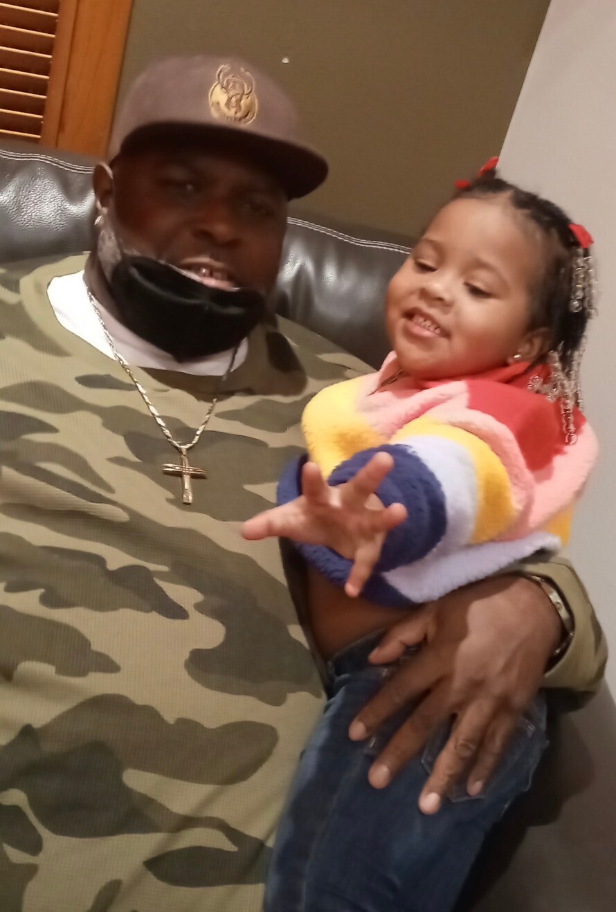 Man in camouflage shirt and cap holding a smiling little girl in a colorful sweater on his lap on a couch.