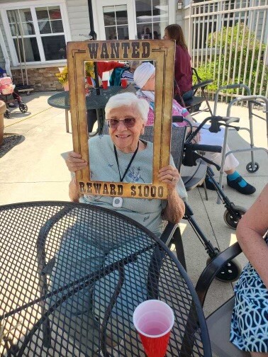 Smiling elderly woman wearing sunglasses holds a wooden 'WANTED' frame with 'REWARD $1000' at an outdoor gathering.