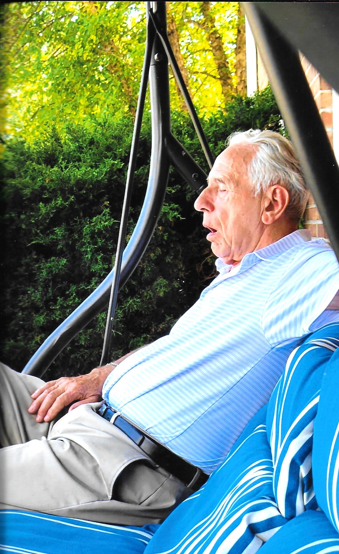 Older man relaxing in a blue-striped polo on a porch swing outdoors, greenery in the background.