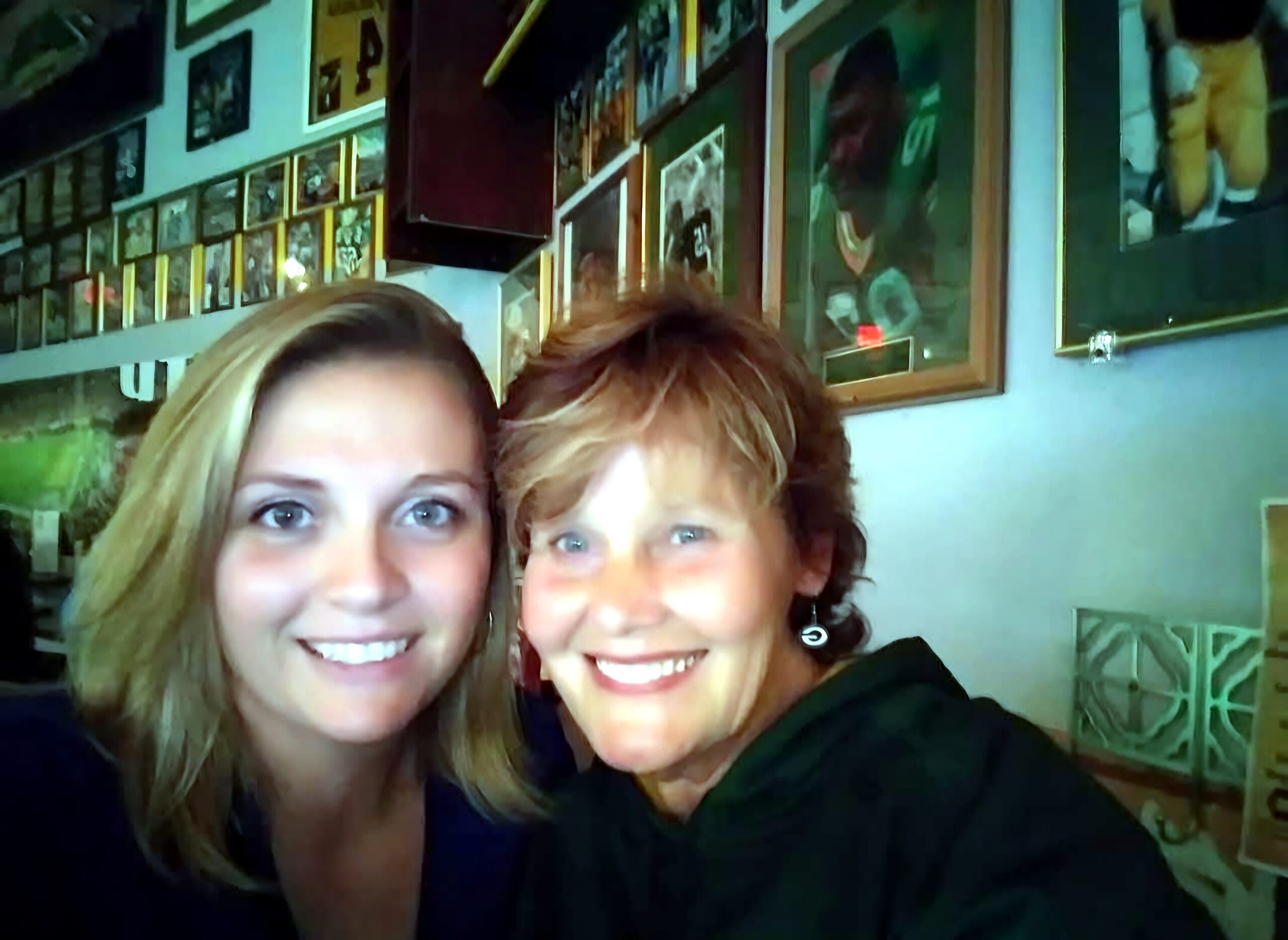 Two smiling women taking a selfie indoors with walls full of framed pictures in the background, creating a cozy, gallery-like setting.