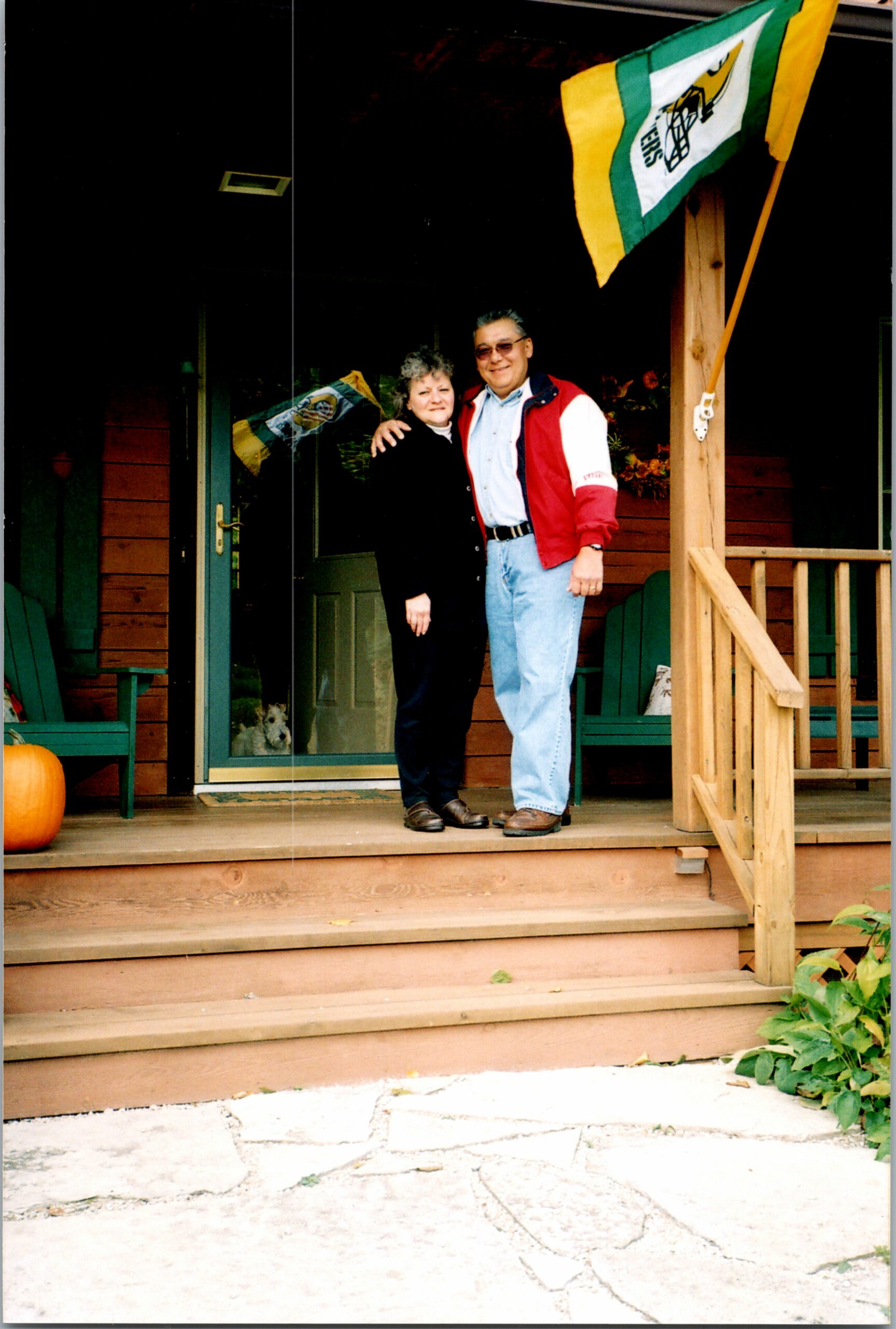 A couple stands together on a wooden porch, smiling, with a green-yellow-white flag in the background and a pumpkin nearby.