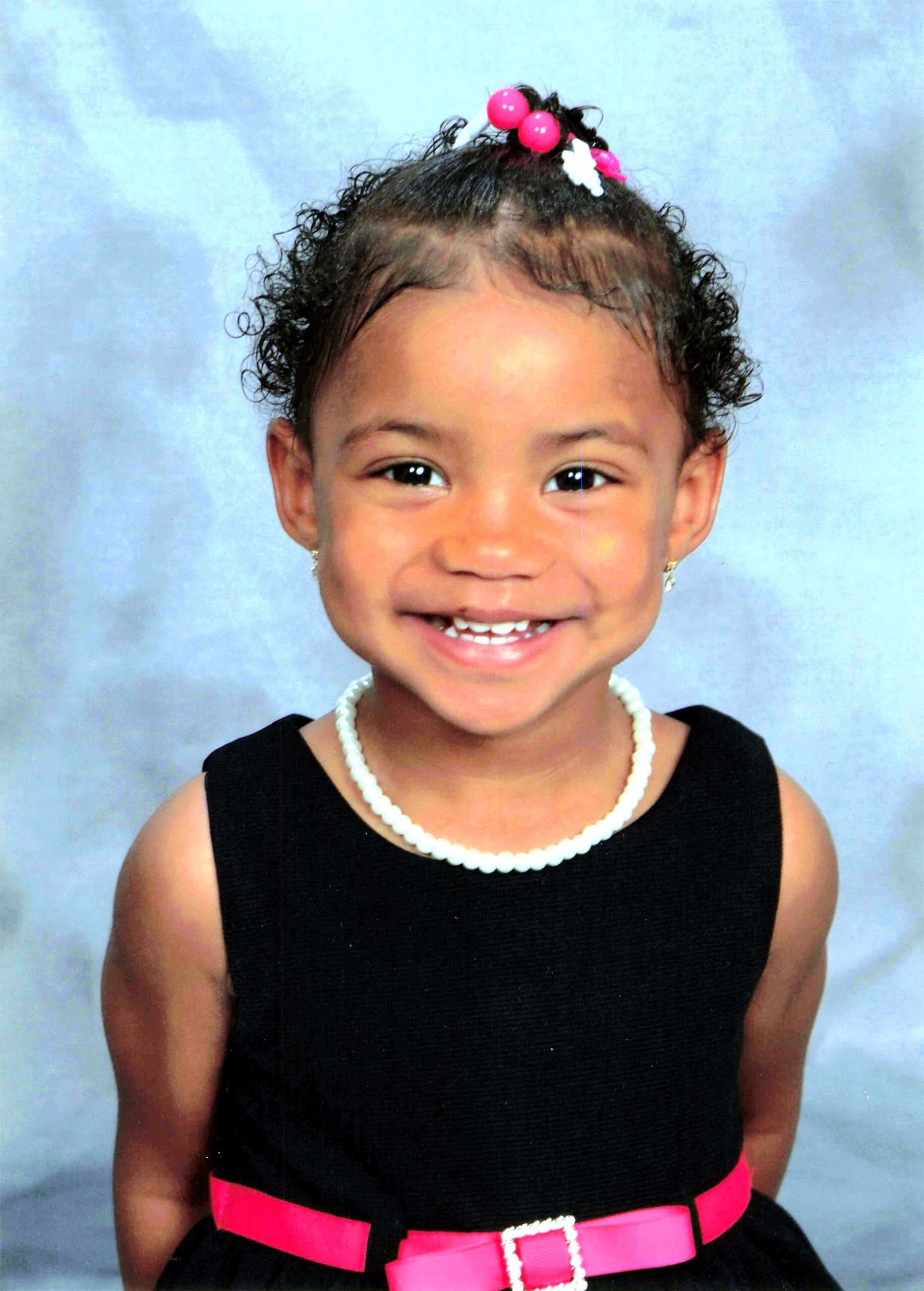 Smiling young girl in a black dress with a pink belt, white pearl necklace, and pink hair accessories against a blue backdrop.