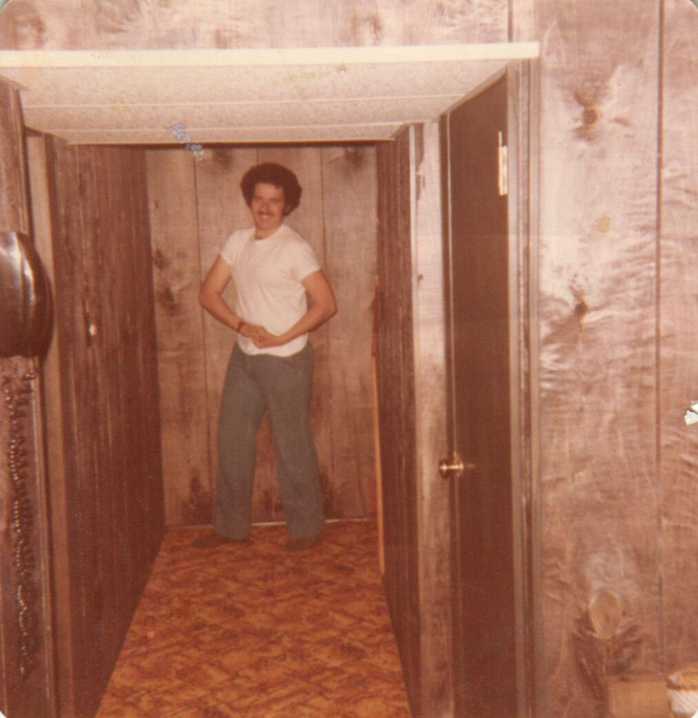 Man in a white tee and jeans flexing his biceps in a narrow wood-paneled hallway, retro setting.