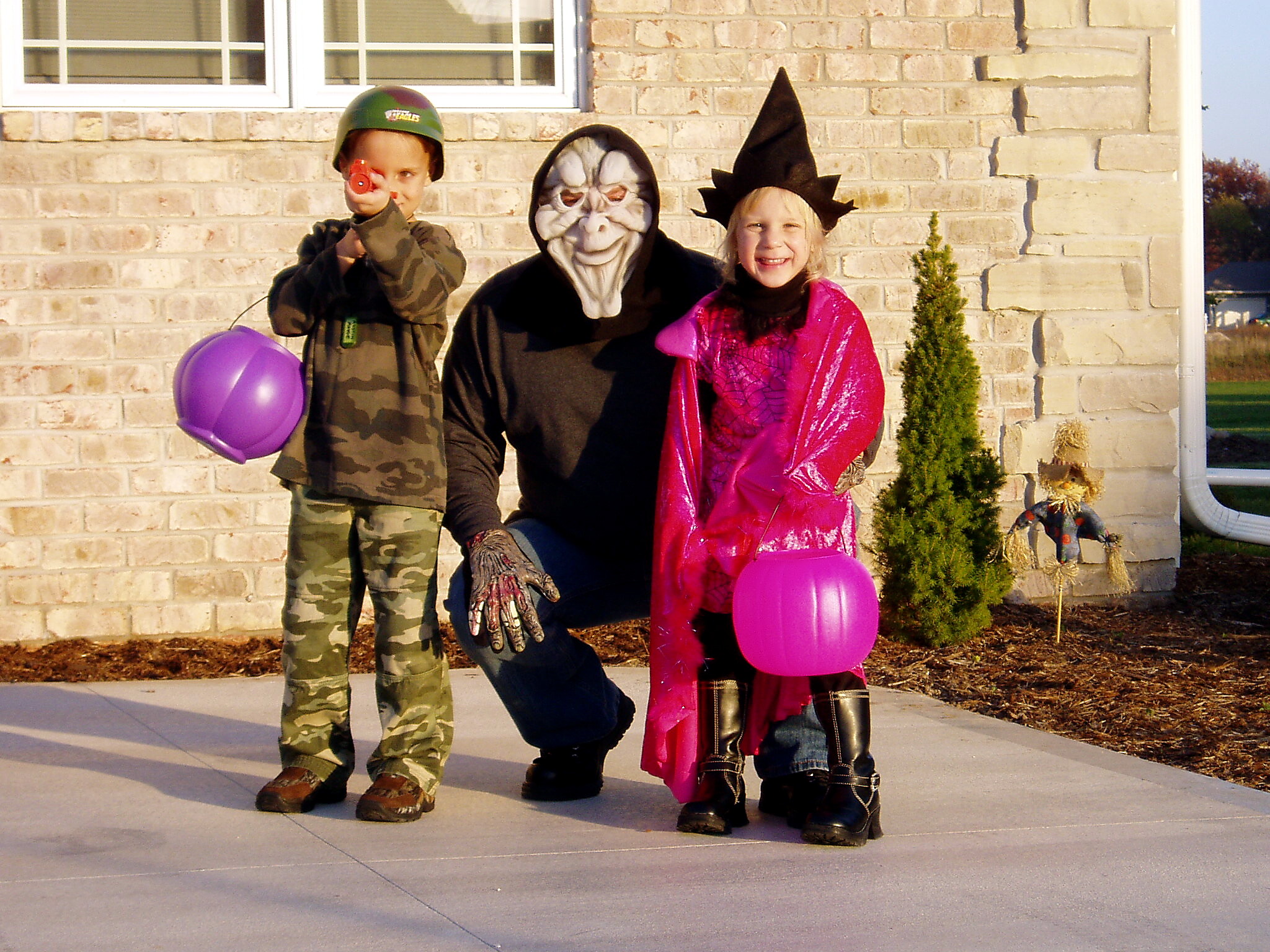 Three kids in Halloween costumes pose outside a brick house; one in camo with helmet, one in a plague doctor mask, and a girl in a pink witch costume, each holding a purple pumpkin.