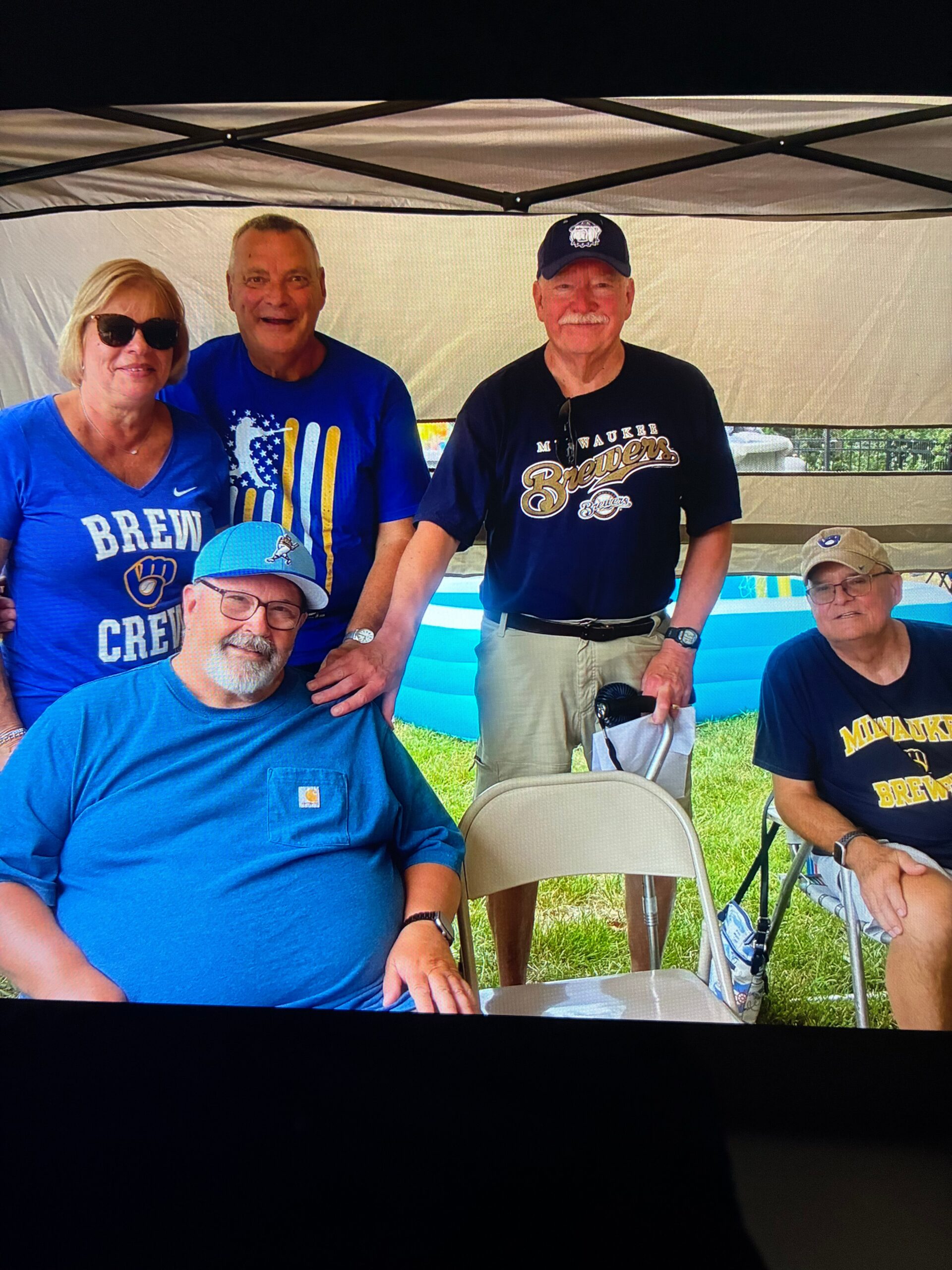 Group of older adults posing under a canopy at an outdoor event; several wear Milwaukee Brewers apparel, including blue shirts and a cap.