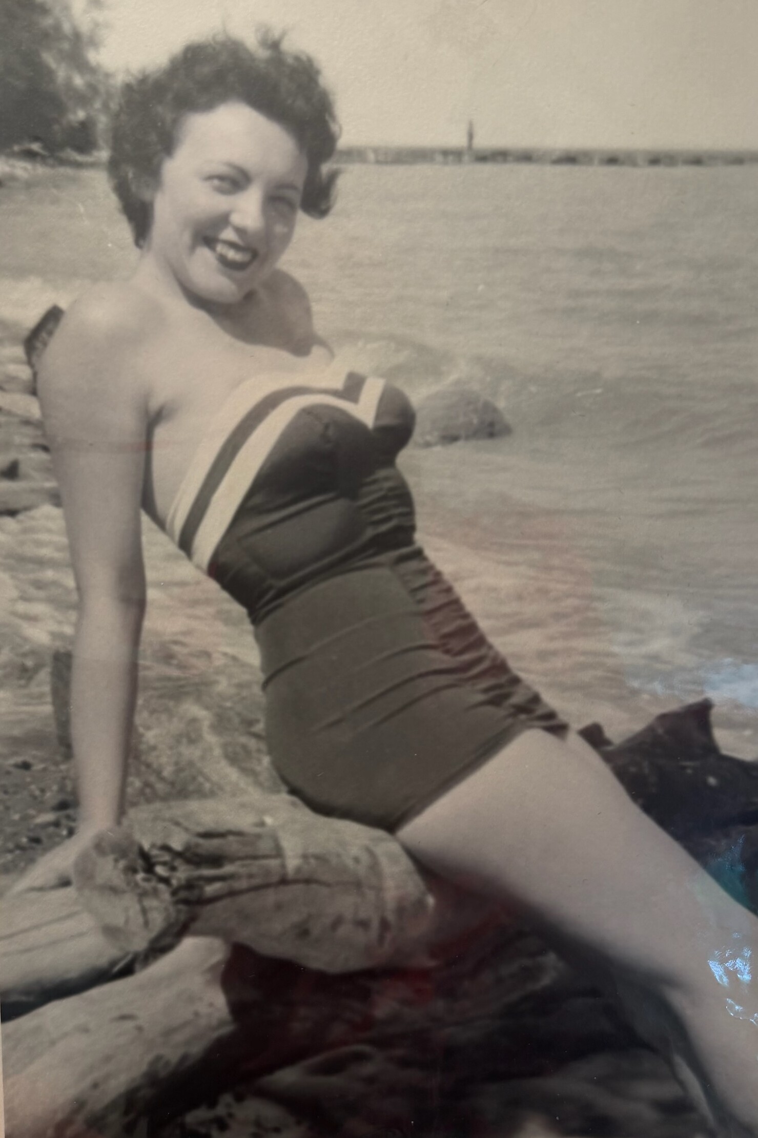 Smiling woman in a vintage one-piece swimsuit leaning on rocks at the beach with the sea in the background (black-and-white photo).