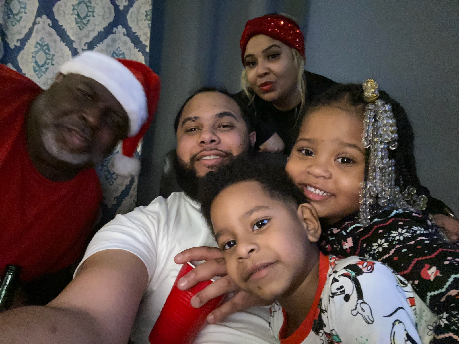 A smiling family of five posing for a Christmas selfie indoors, wearing holiday outfits and Santa-themed accessories (Santa hat, red sequined headband).