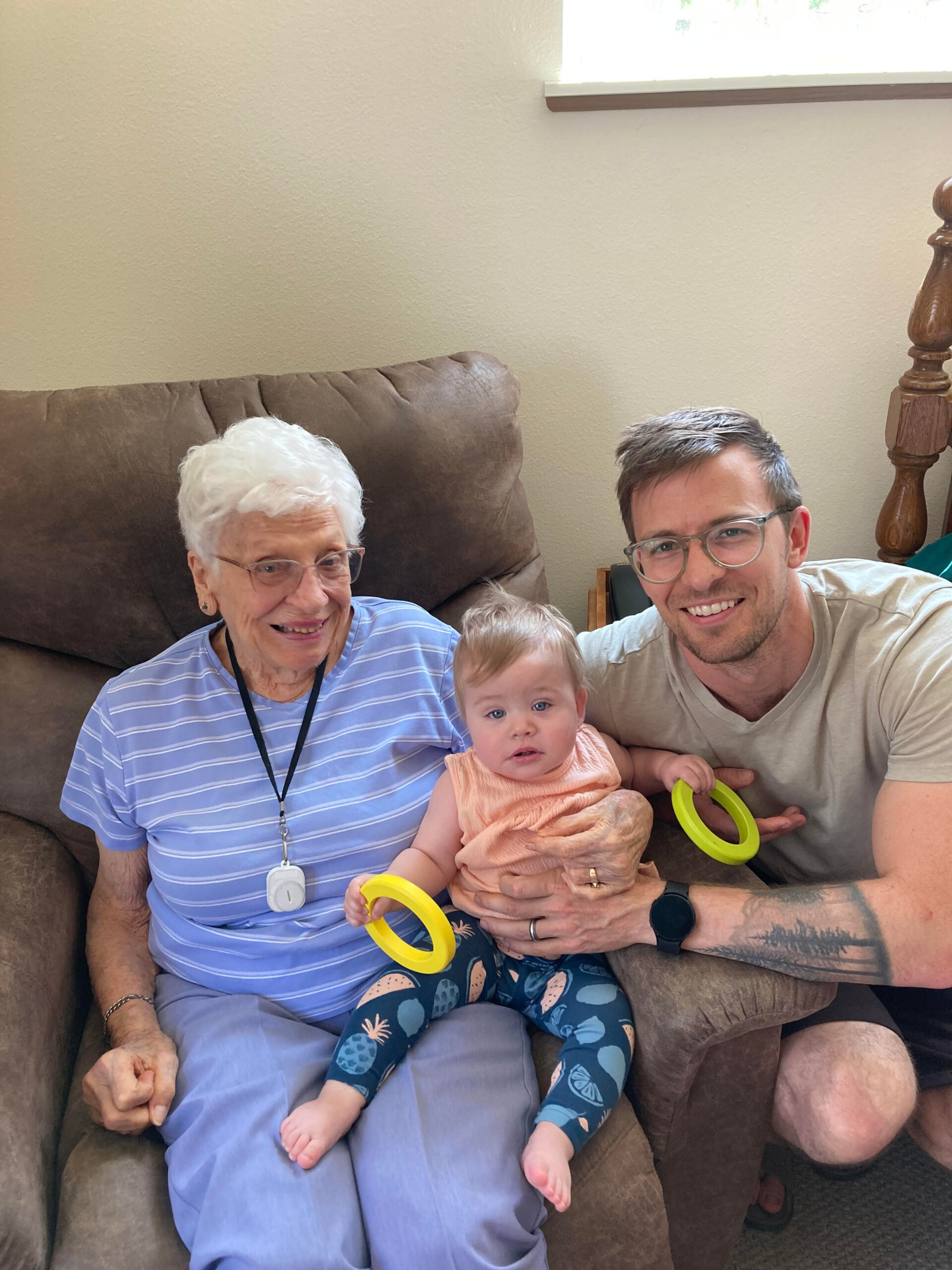 Three generations sit together on a brown sofa: an elderly woman with white hair, a baby in a peach outfit, and a smiling man with glasses, holding bright yellow teething rings indoors.