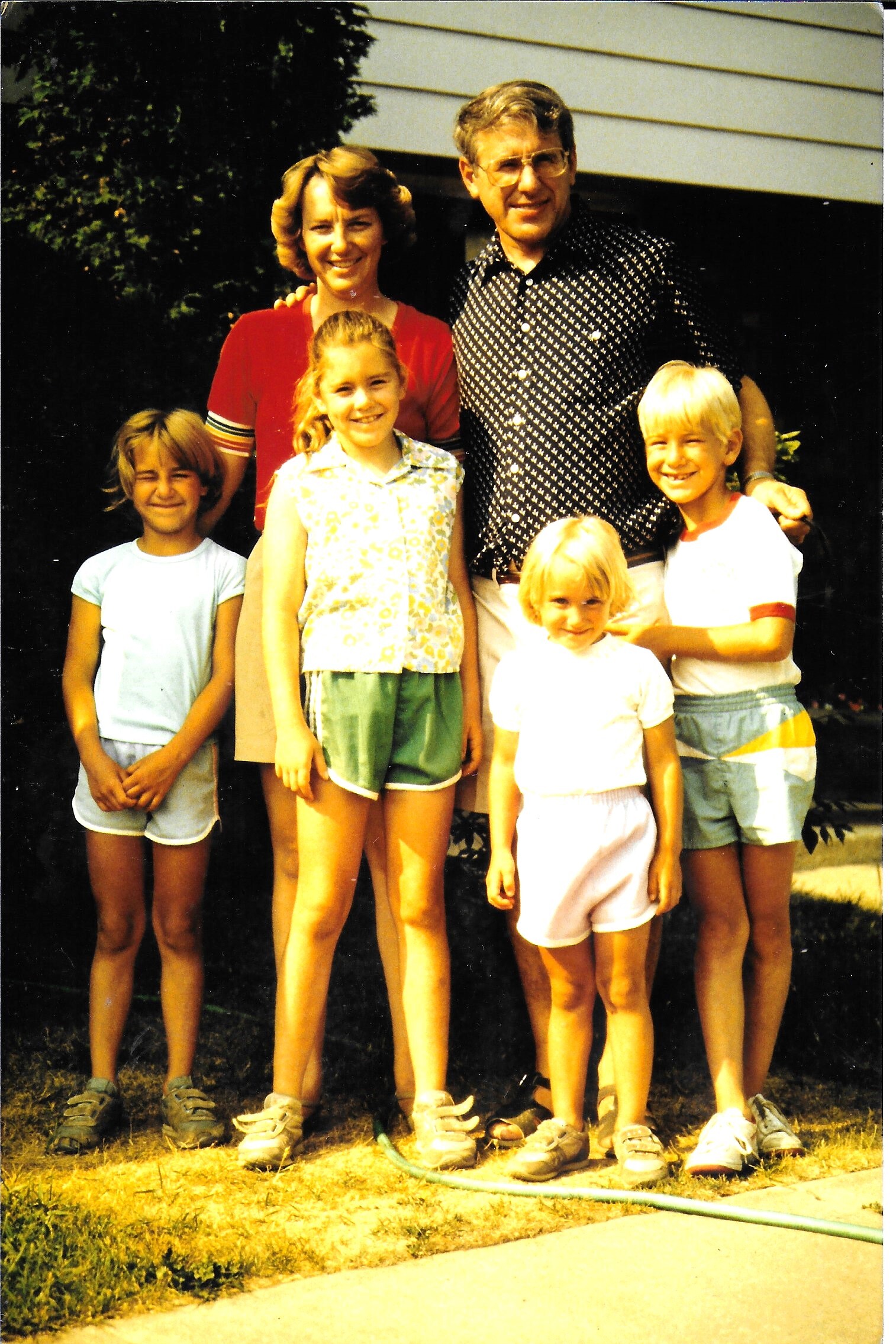 Smiling family of six posing outdoors in front of a house; two adults behind four children in summer clothing.