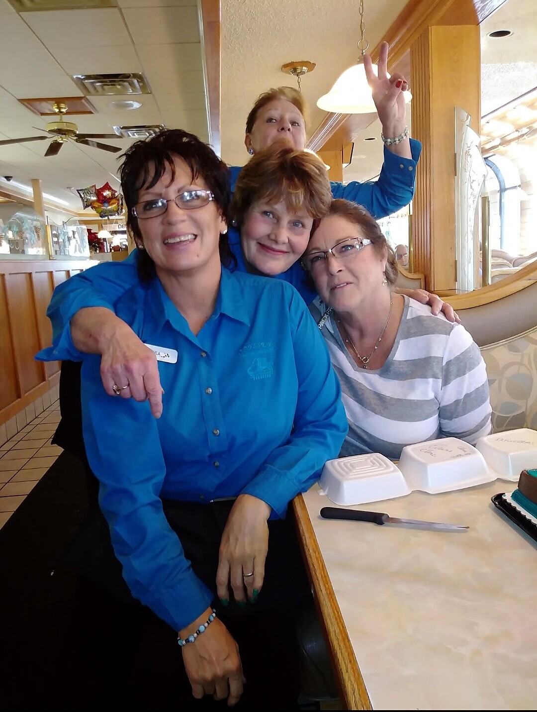 Three women sit at a diner table, smiling for a group photo; one in a blue shirt with a name tag sits front left as others lean in from behind and side.