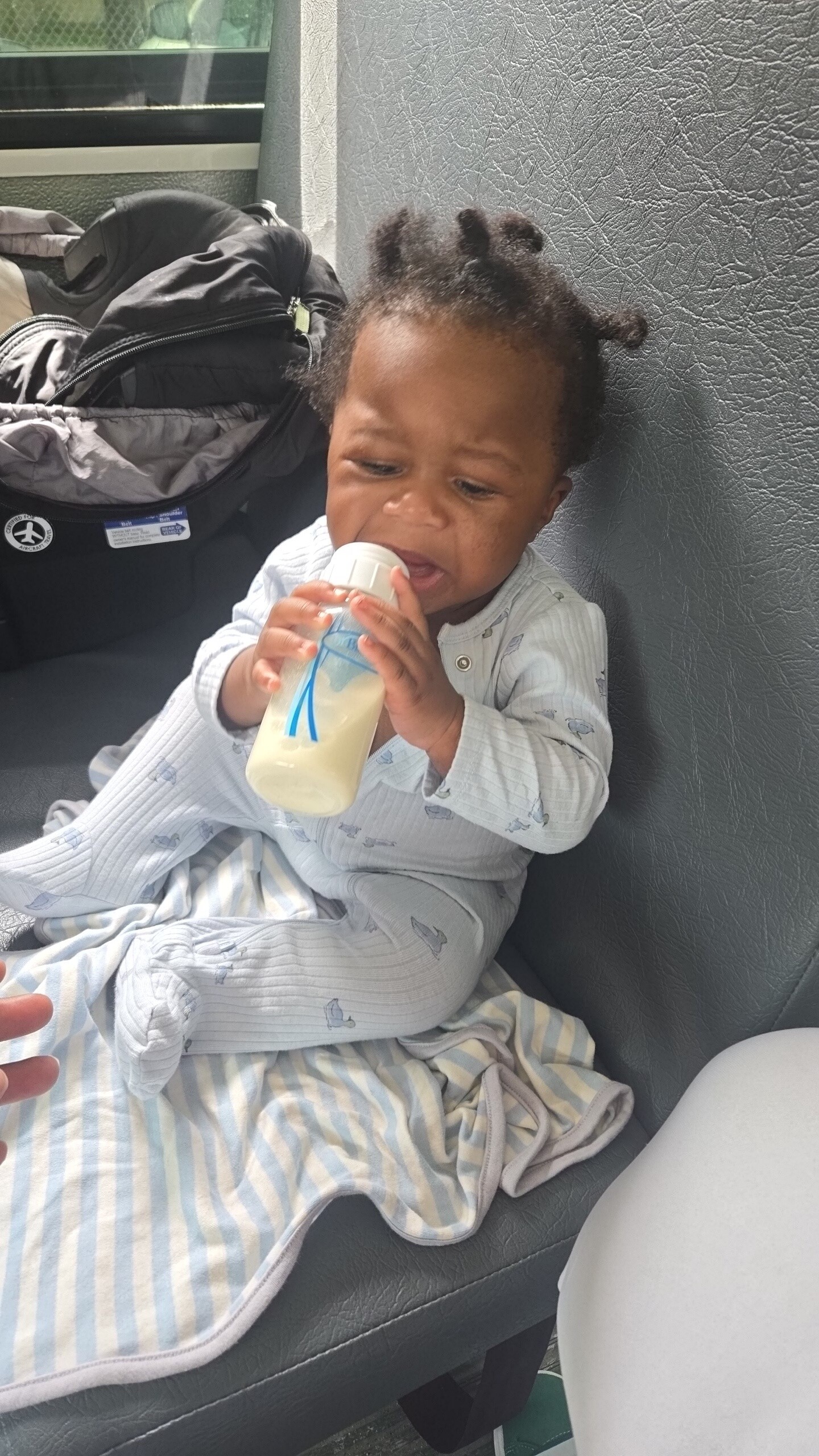 Toddler in light blue striped pajamas drinks from a baby bottle while sitting on a car/bus seat, with a backpack nearby