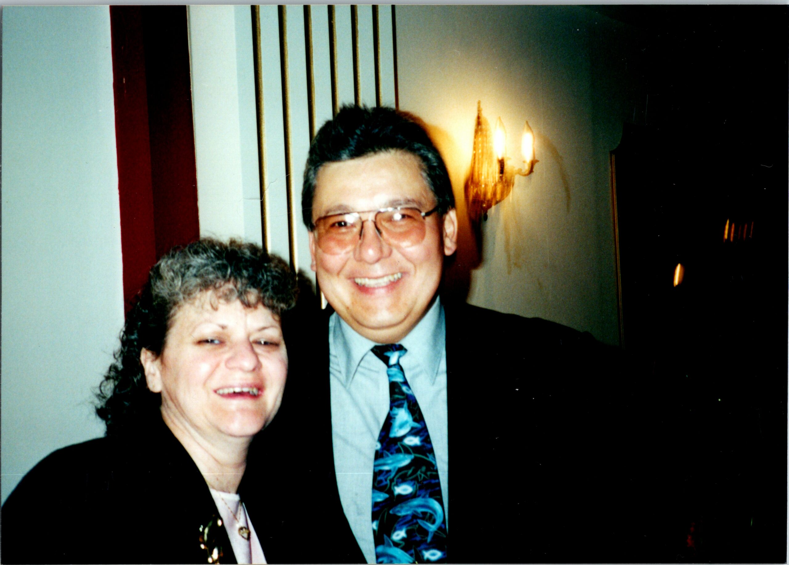 Smiling man in glasses and a patterned tie stands beside a woman in formal attire at an indoor event.