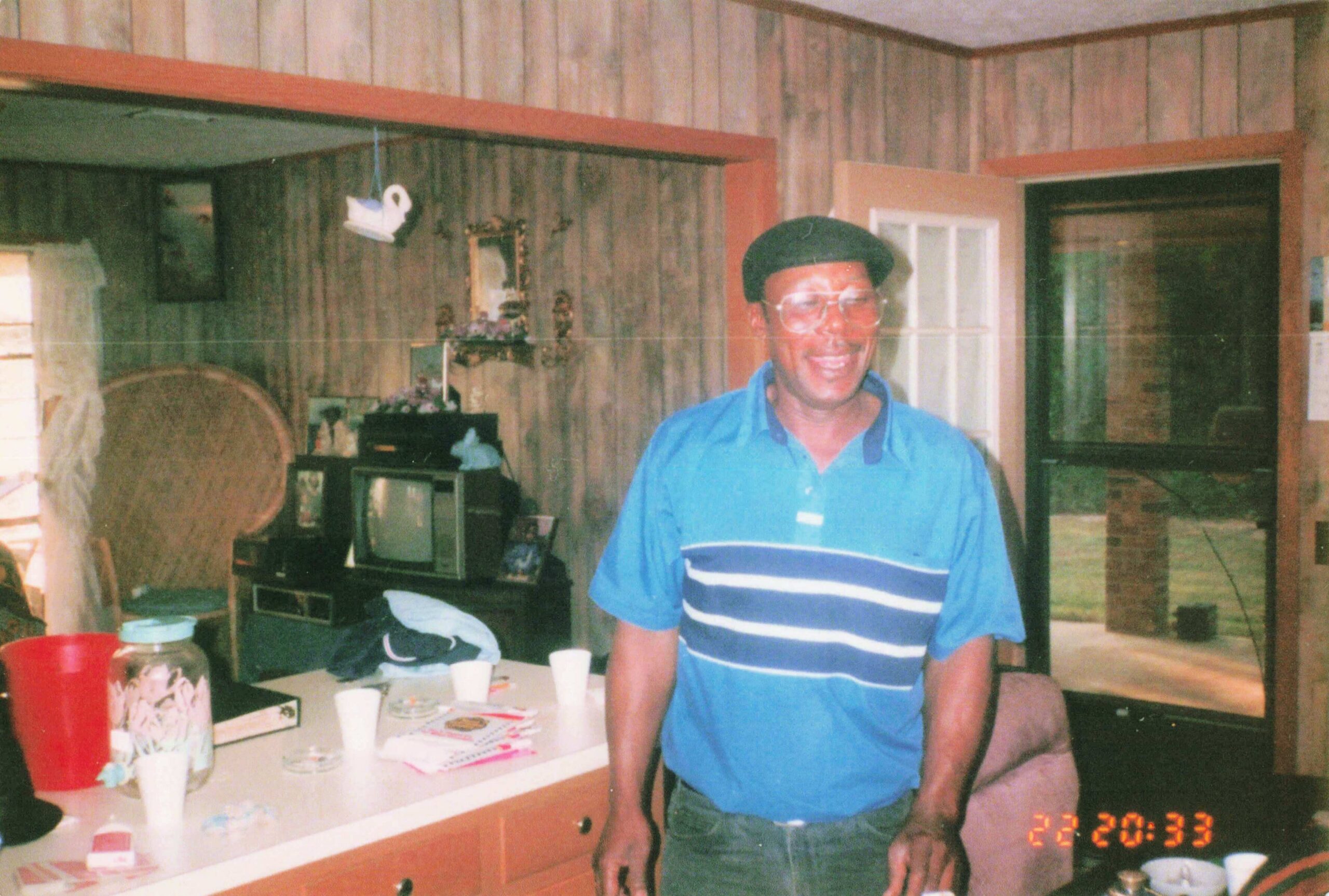 Smiling man in a blue polo shirt and glasses stands by a cluttered kitchen counter with paper cups and snacks; wood-paneled walls surround him.
