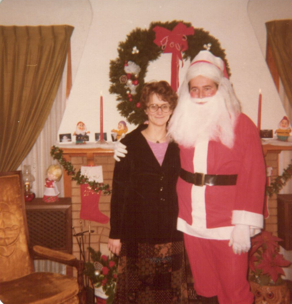 Woman with glasses posing next to a man dressed as Santa Claus in a Christmas‑themed living room with a wreath and stockings on the mantelpiece.