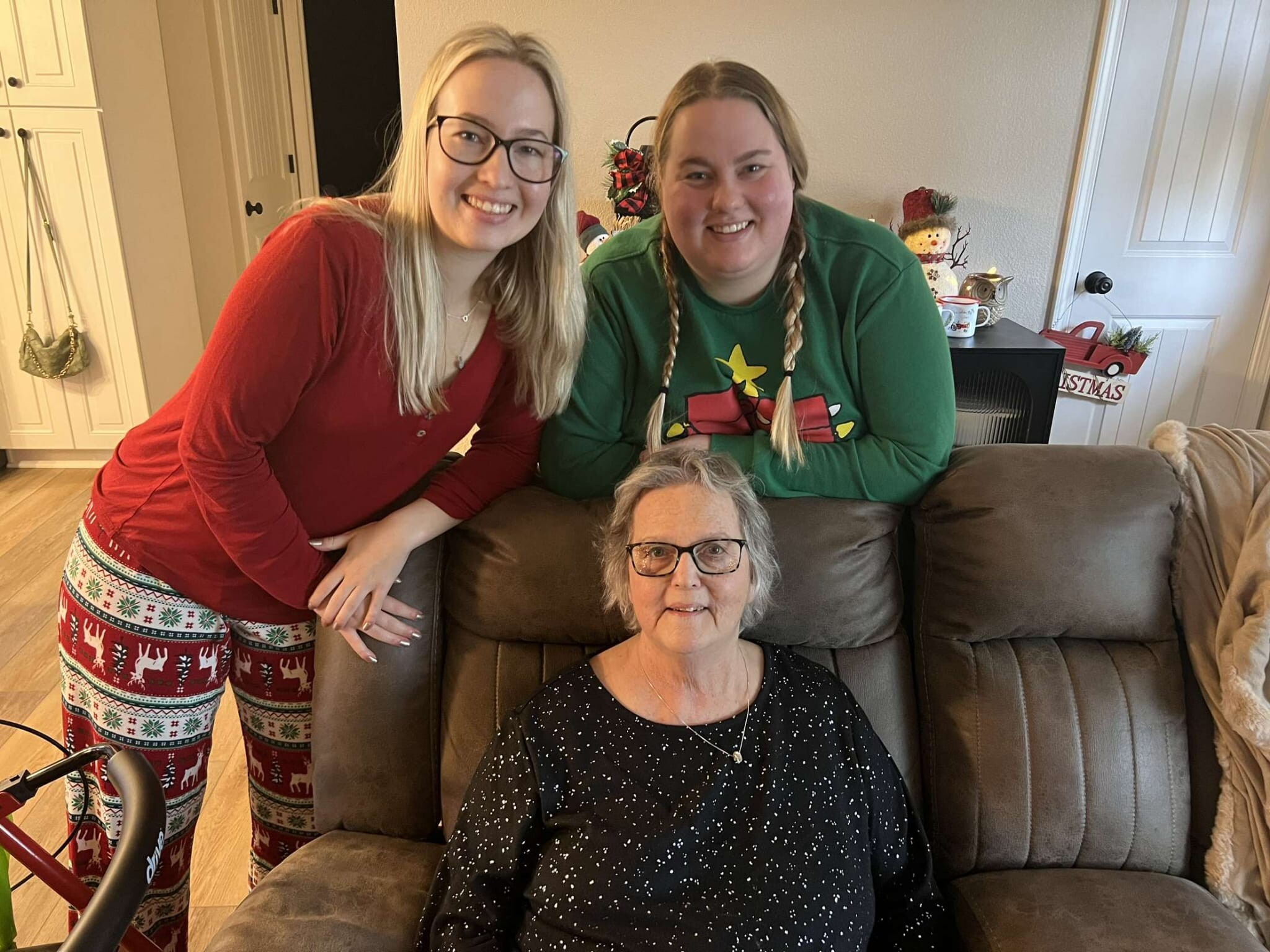 Three women pose together in a cozy living room holiday scene: an older woman with glasses sits on a brown couch while two others stand beside her, all smiling. Christmas attire and decorations are visible in the background.