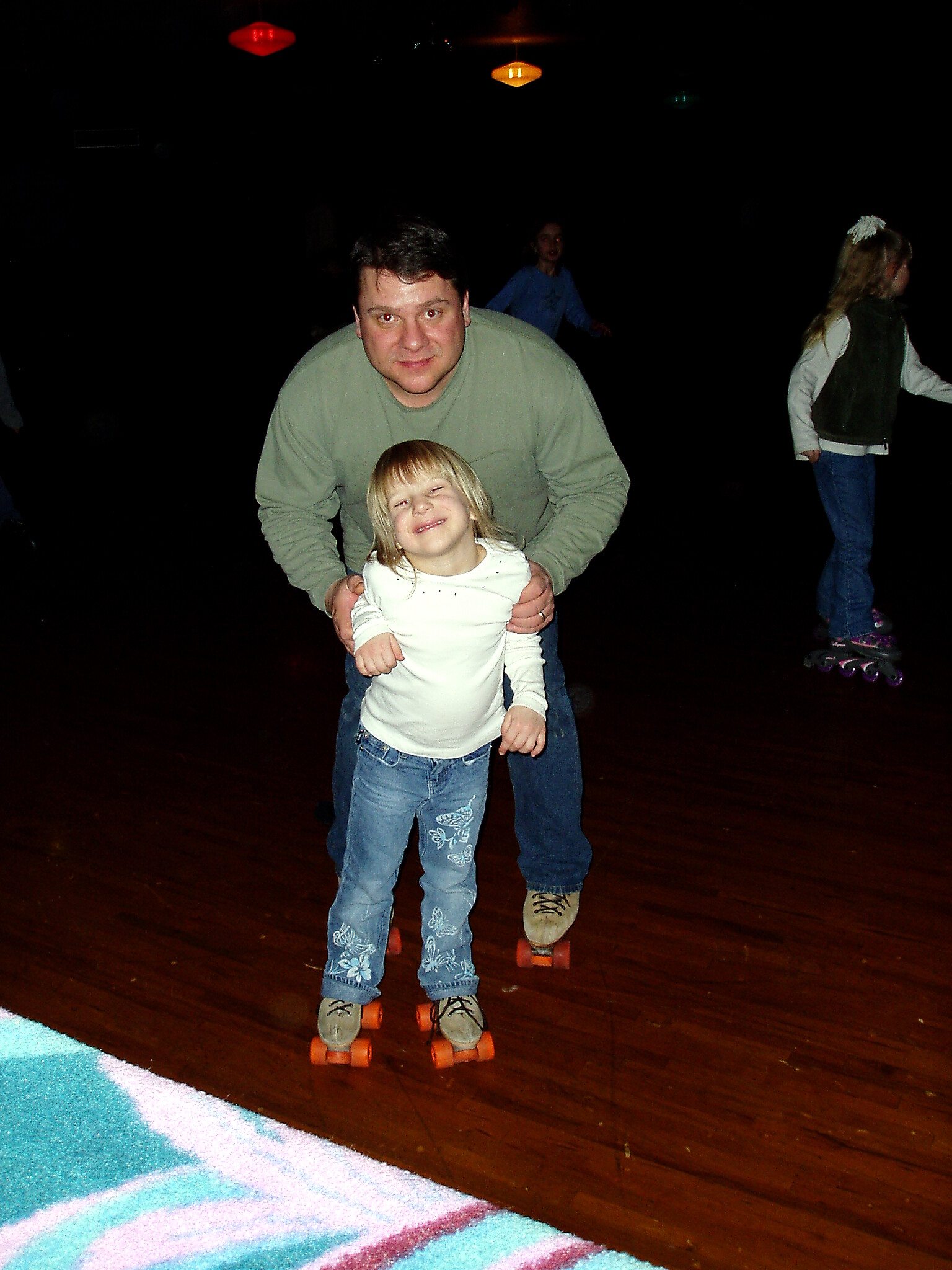 Man gently helping a smiling young girl on orange roller skates in a dim indoor roller rink.