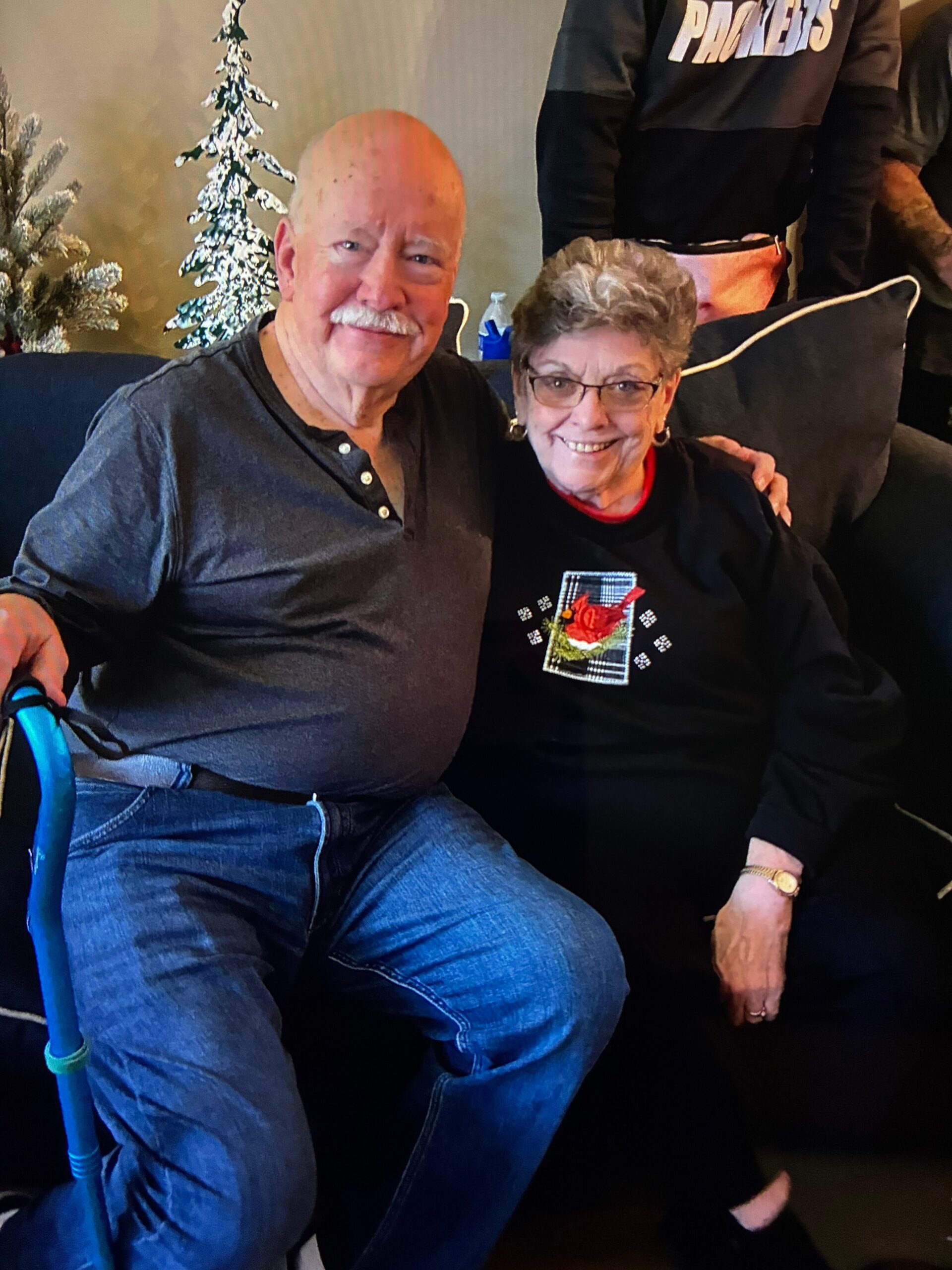 Smiling elderly couple seated on a sofa; man in a dark shirt and jeans, woman in a black top with a decorative patch, holiday trees behind them.