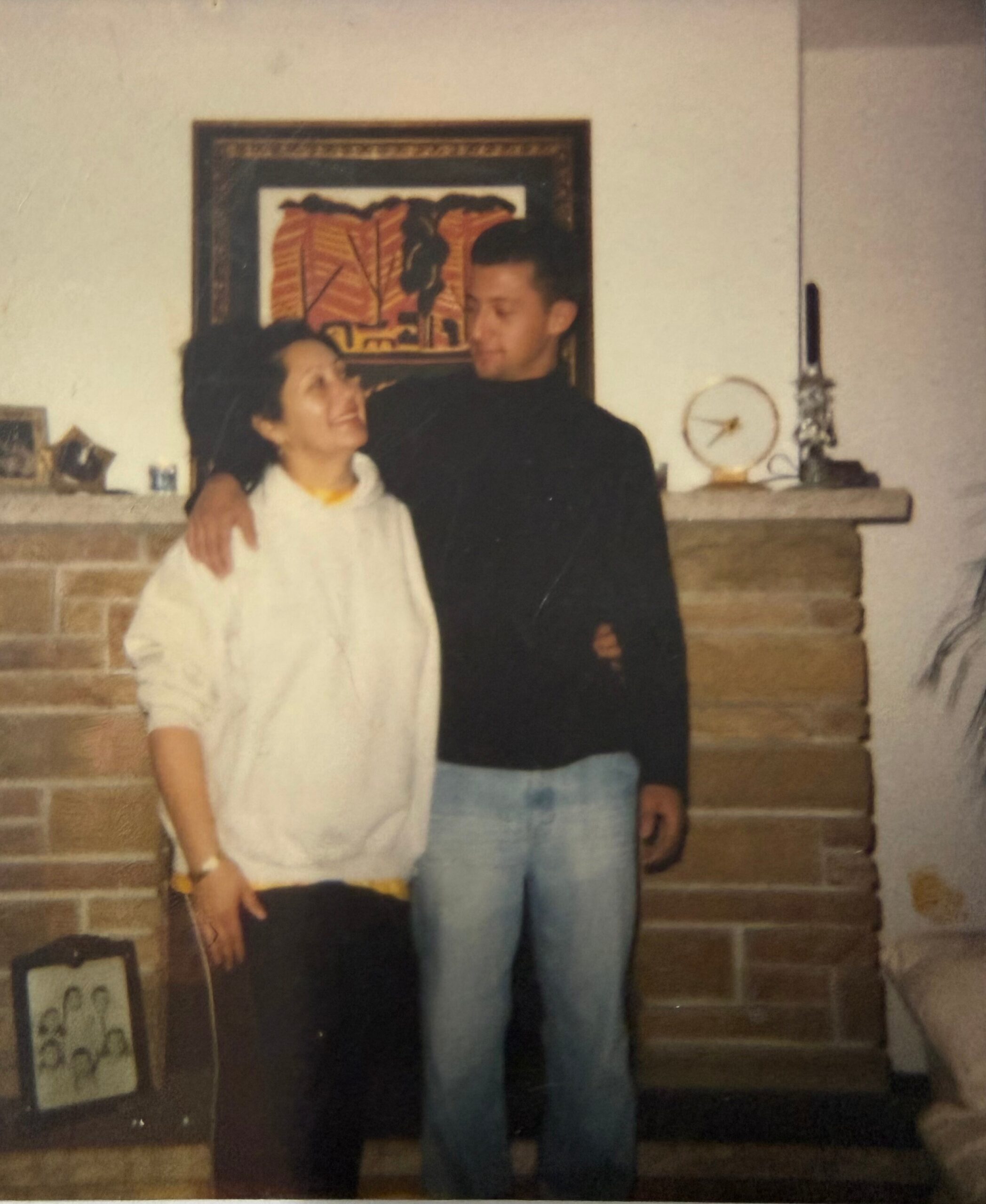 Two friends stand close together in a living room, one with an arm around the other, smiling by a brick fireplace and mantle decor behind them and a framed art piece above.