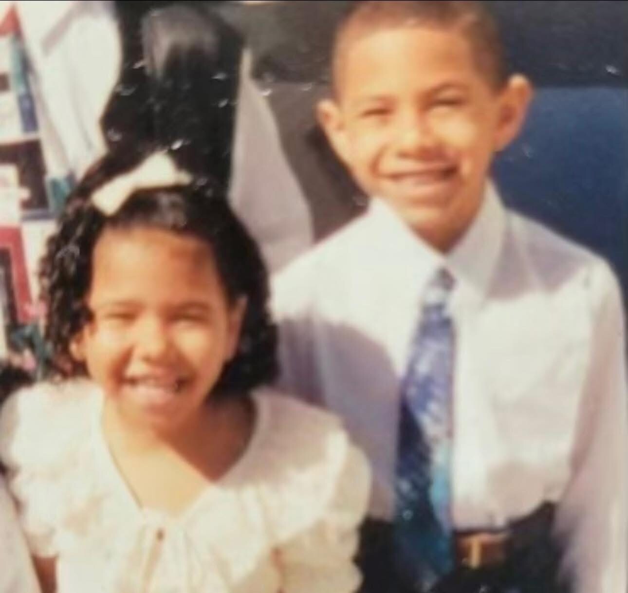 Two smiling children posing together for a photo, girl on the left and a boy in a white shirt and blue patterned tie on the right.