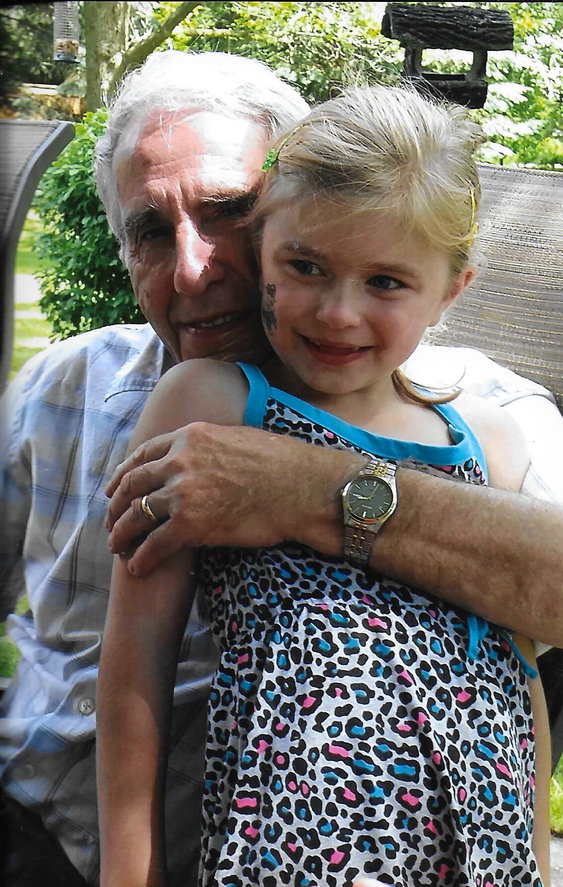 An older man with gray hair hugs a smiling young girl outdoors, seated in a garden.