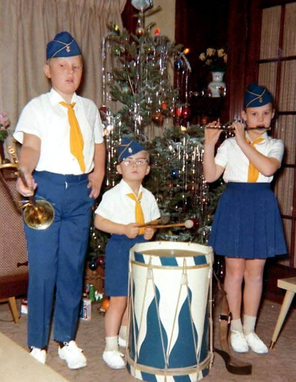 Three children in matching white shirts, blue skirts/ shorts and yellow ties playing musical instruments by a Christmas tree: trumpet, snare drum, and flute.