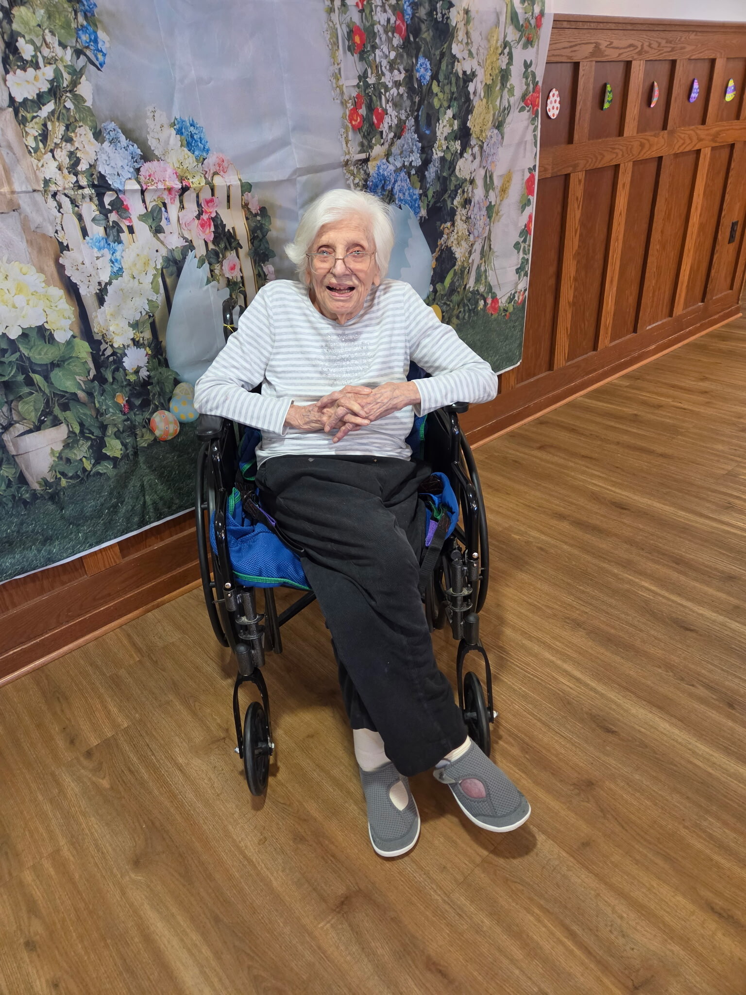 Smiling elderly woman in a wheelchair, wearing a light striped shirt, with hands folded in her lap, in front of a floral backdrop in a wooden-floored room.