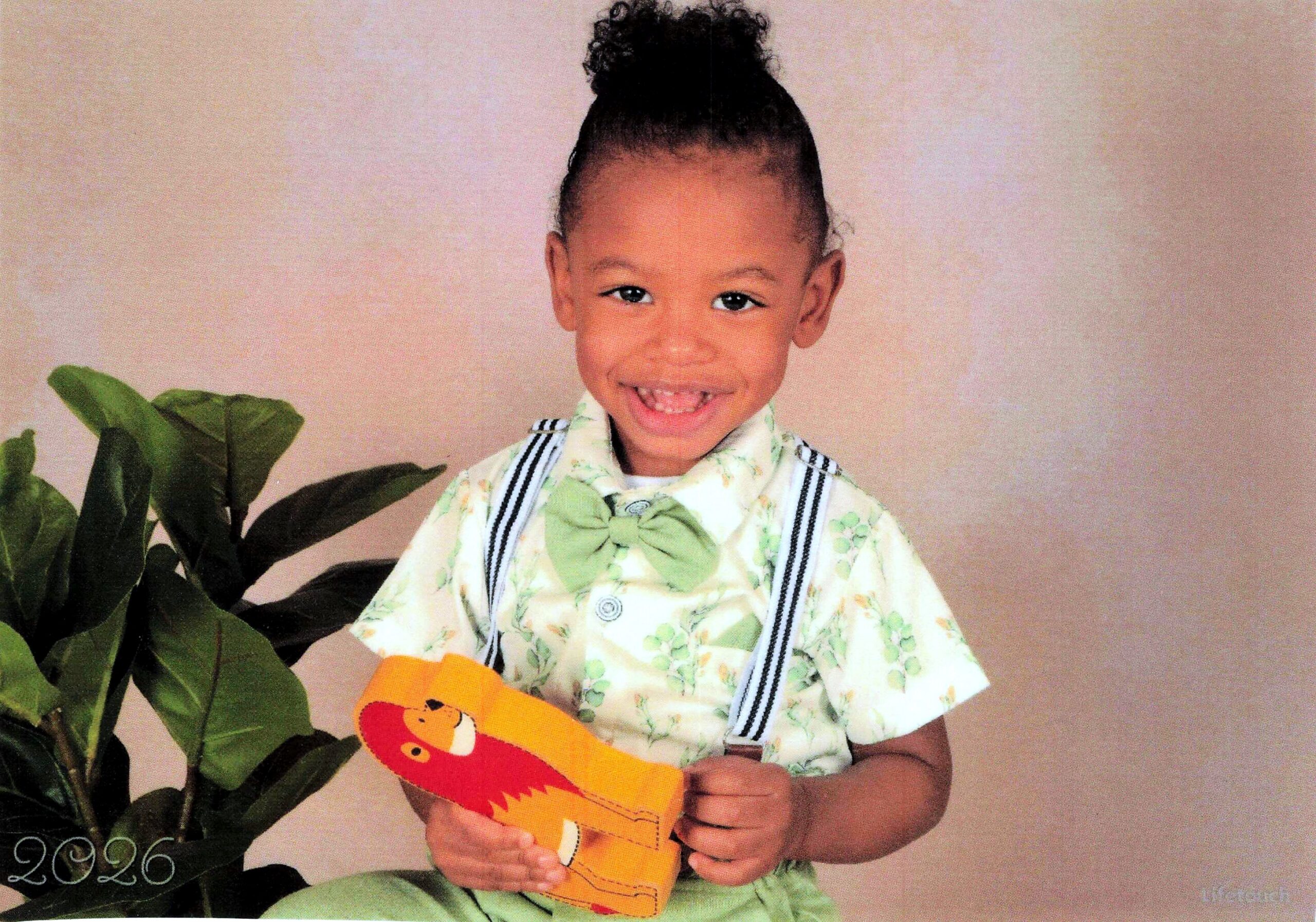Smiling young boy in a floral shirt with a green bow, holding an orange crocodile toy in front of a plant and beige background.