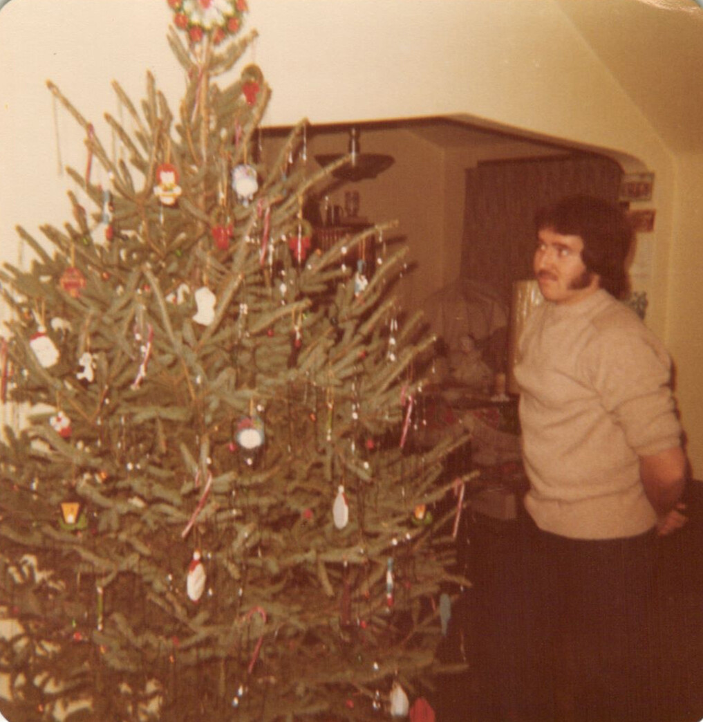 Man in a beige sweater stands beside a decorated Christmas tree in a living room interior.
