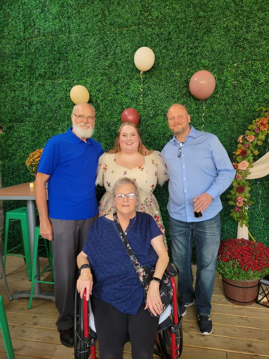 Four people posing at a celebration: an elderly woman in a wheelchair sits front and center, flanked by two men and a woman against a green hedge with balloons behind them on a wooden deck.