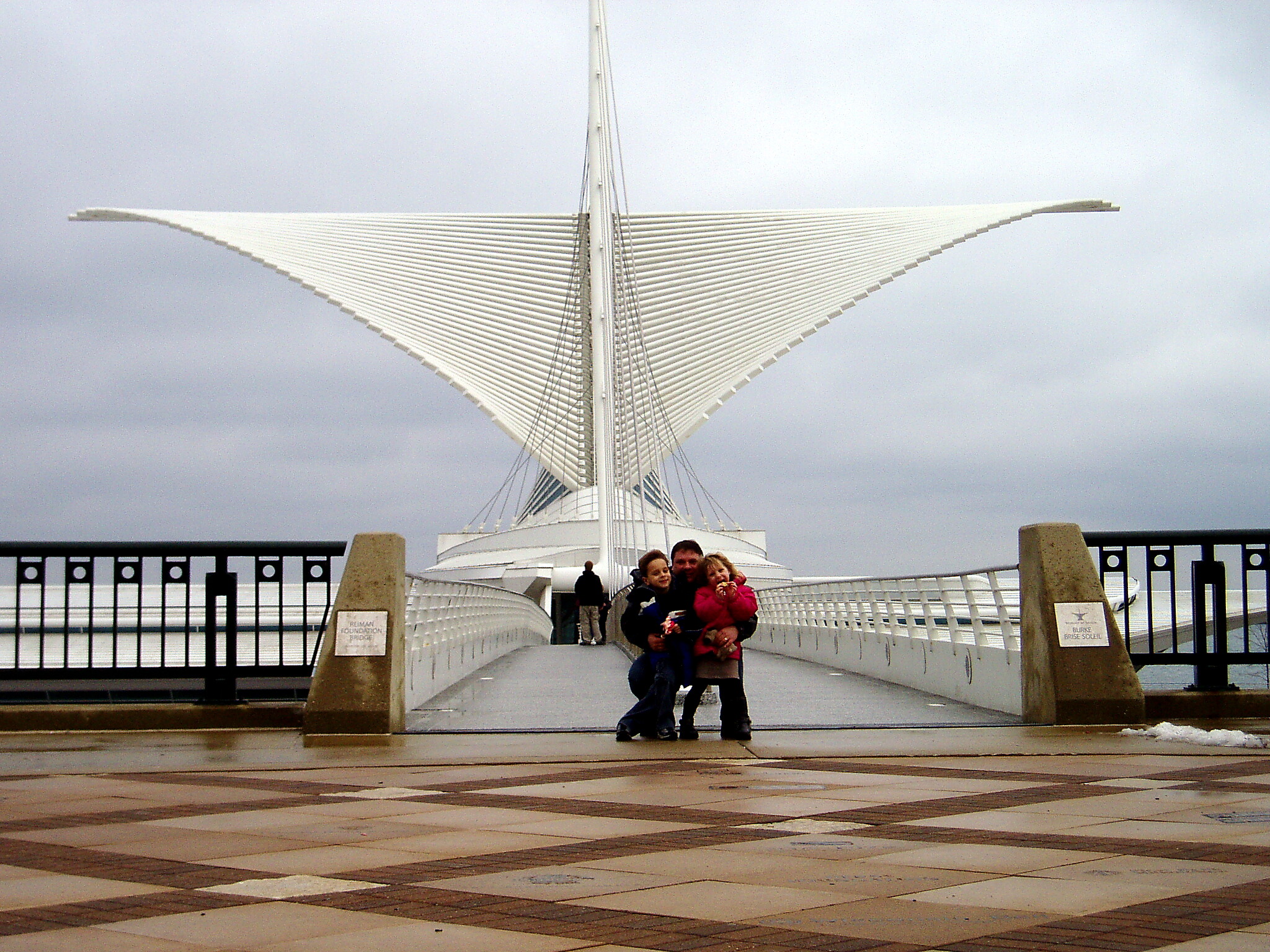 Group of people posing for a photo on a modern white bridge with a tall central pylon and sweeping cable arches against a cloudy sky.