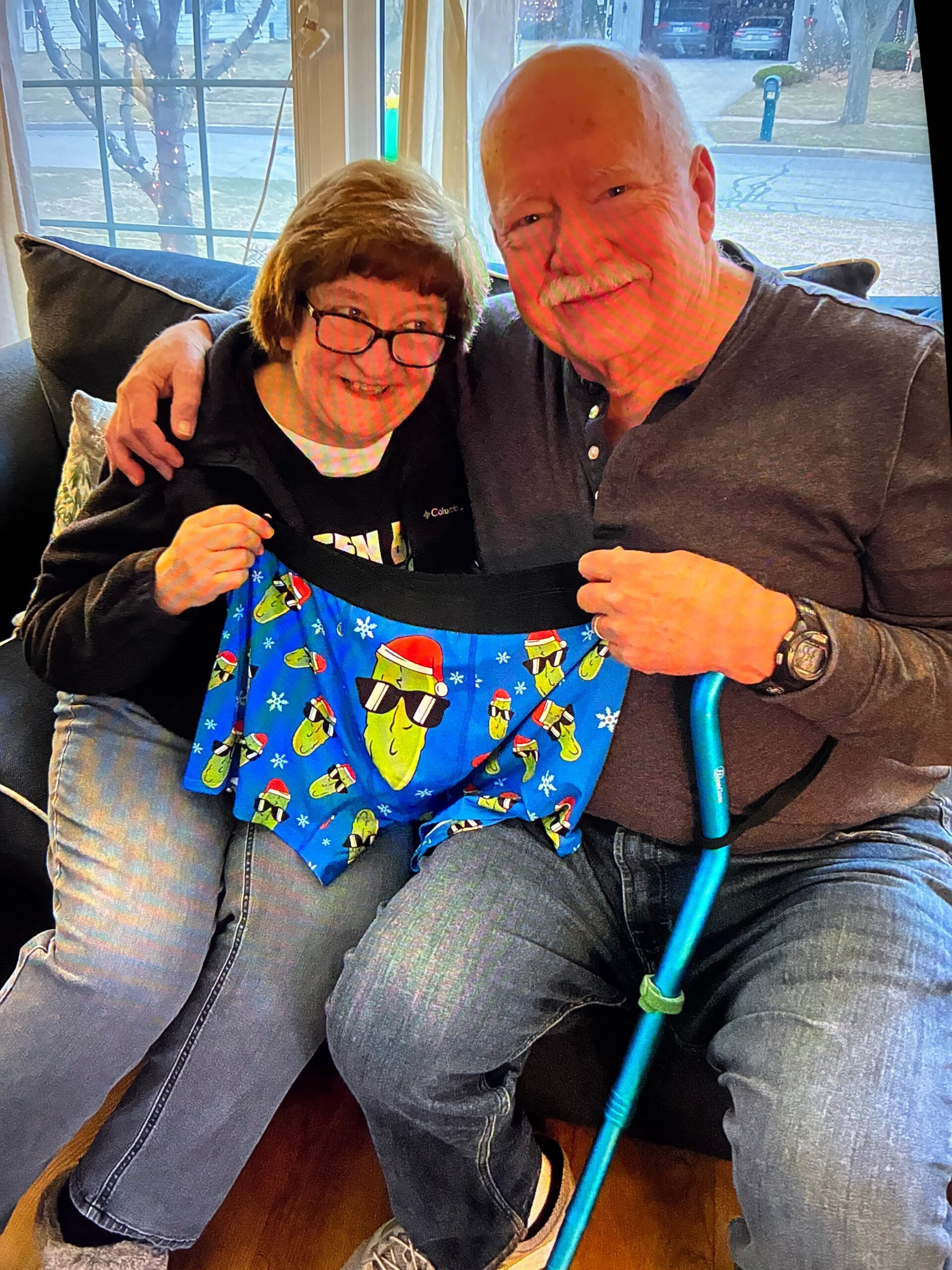 Smiling elderly couple sitting close on a sofa indoors; woman holds a blue holiday gift bag while the man rests his arm around her.
