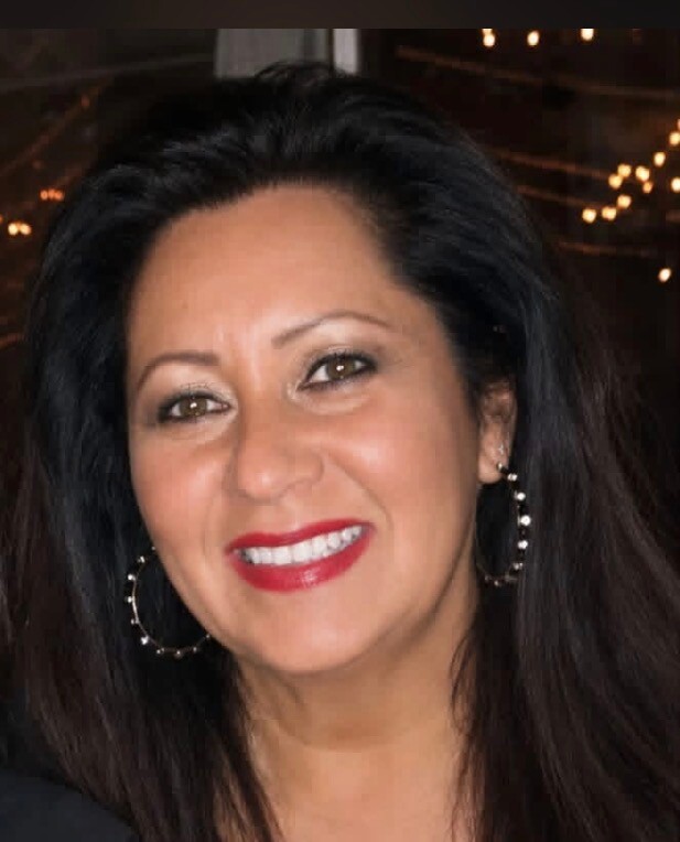 Smiling woman with long dark hair, red lipstick, and large hoop earrings at a nighttime event.