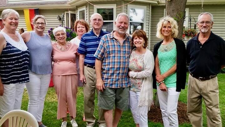 Group of ten adults posing for a photo in a front yard of a house, smiling at the camera on a sunny day.