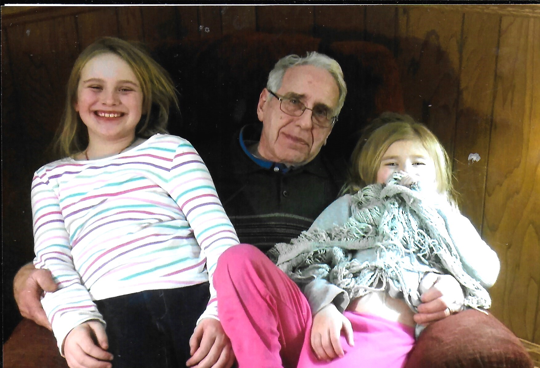 Three generations posing together: an elderly man flanked by two smiling girls on a wooden backdrop.