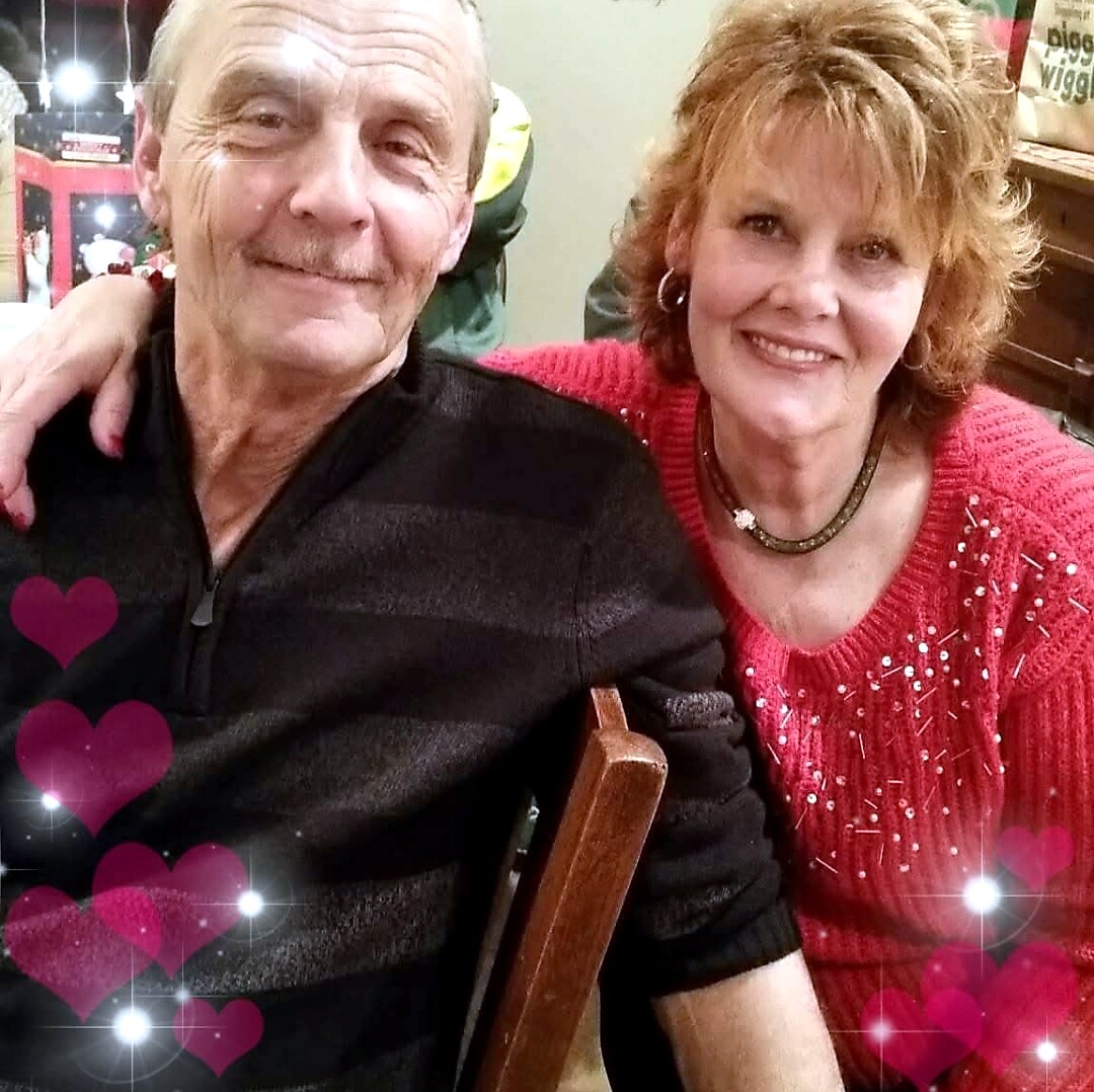 Smiling elderly couple seated together; woman in a red sweater with short hair, man in a dark shirt, pink heart overlays.