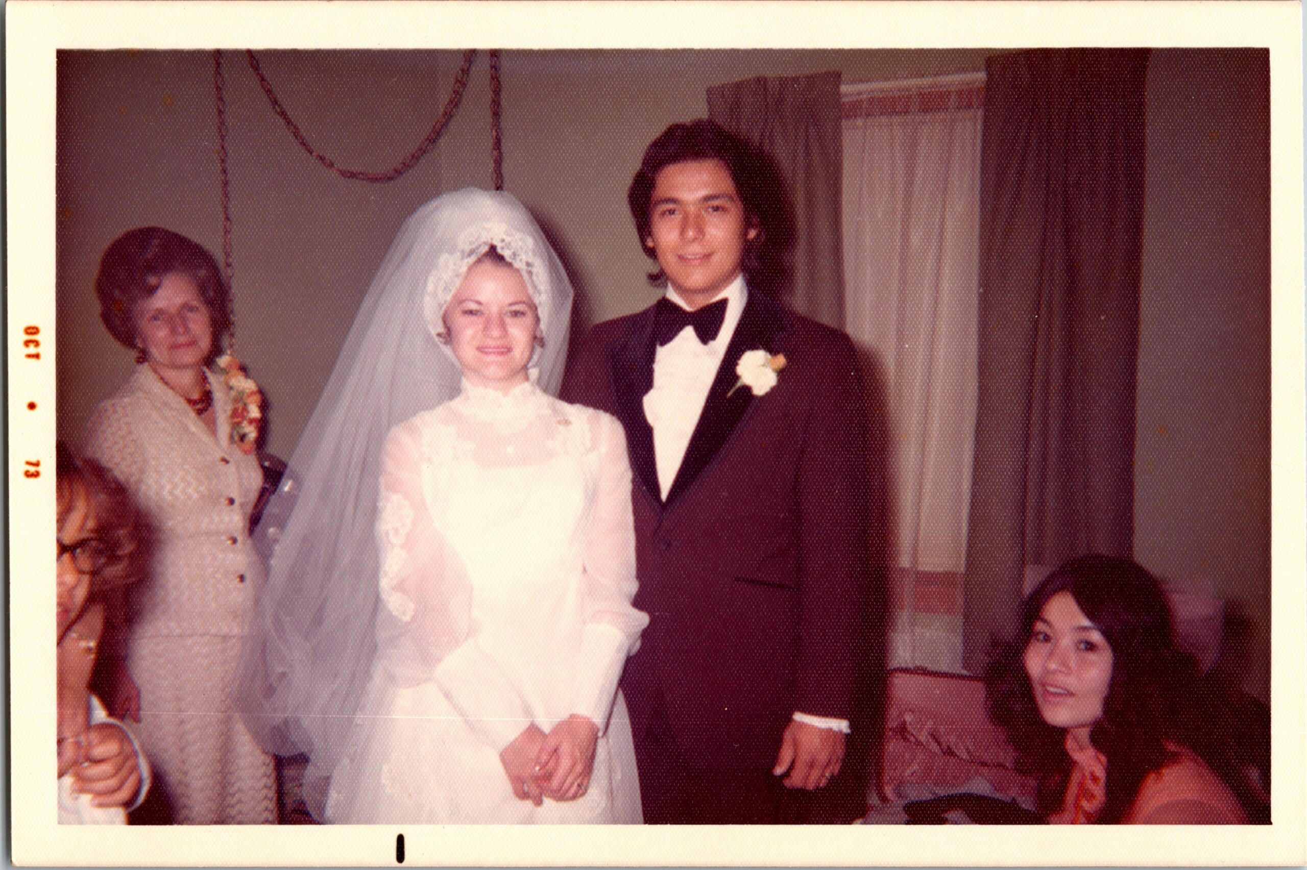 Bride in a lace wedding gown and veil stands with groom in a burgundy tuxedo; a woman with a bouquet watches on the left, guests seated in the background.