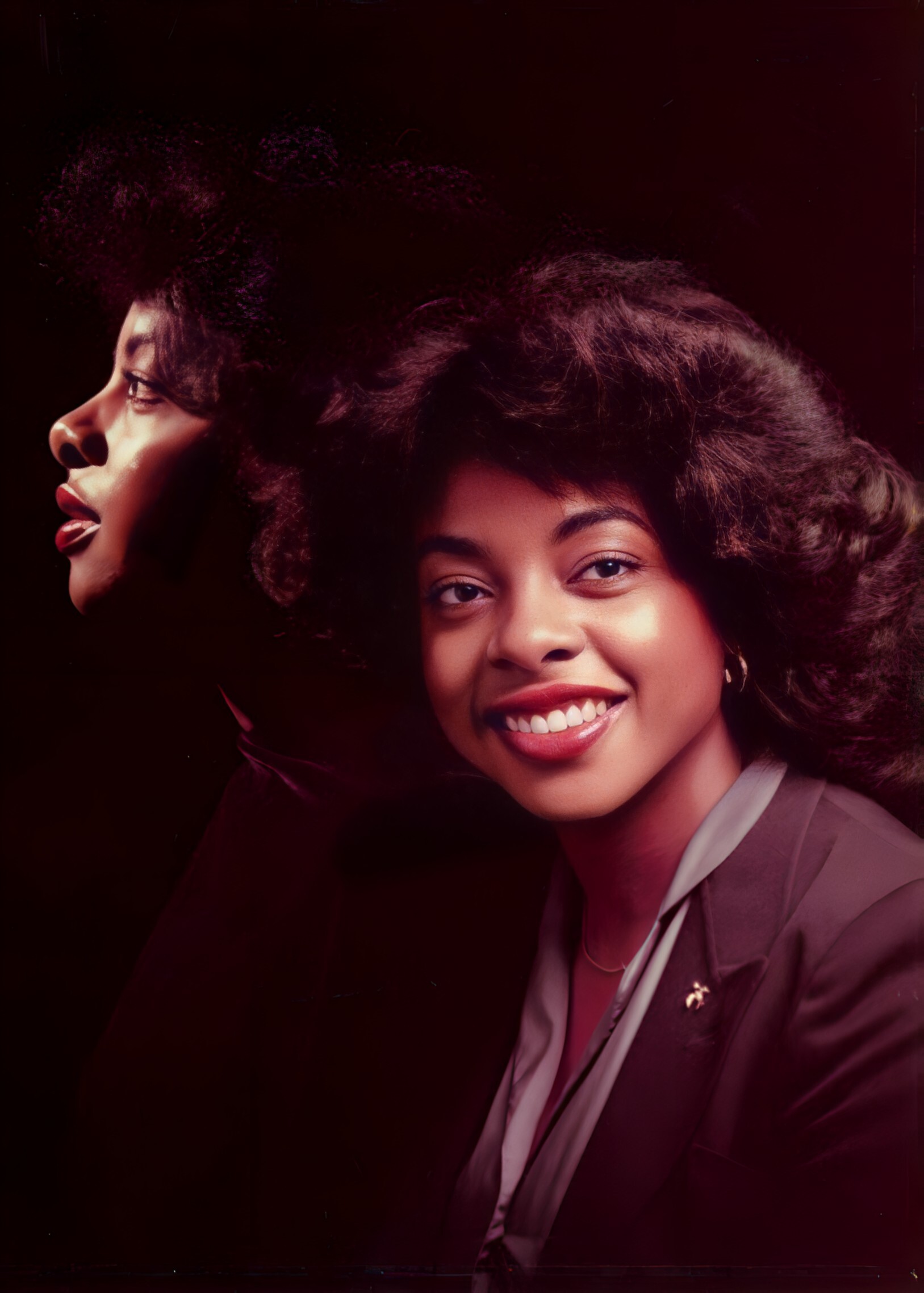 Portrait of two women, one facing left in profile and another smiling toward the camera, with a dark red-toned background.
