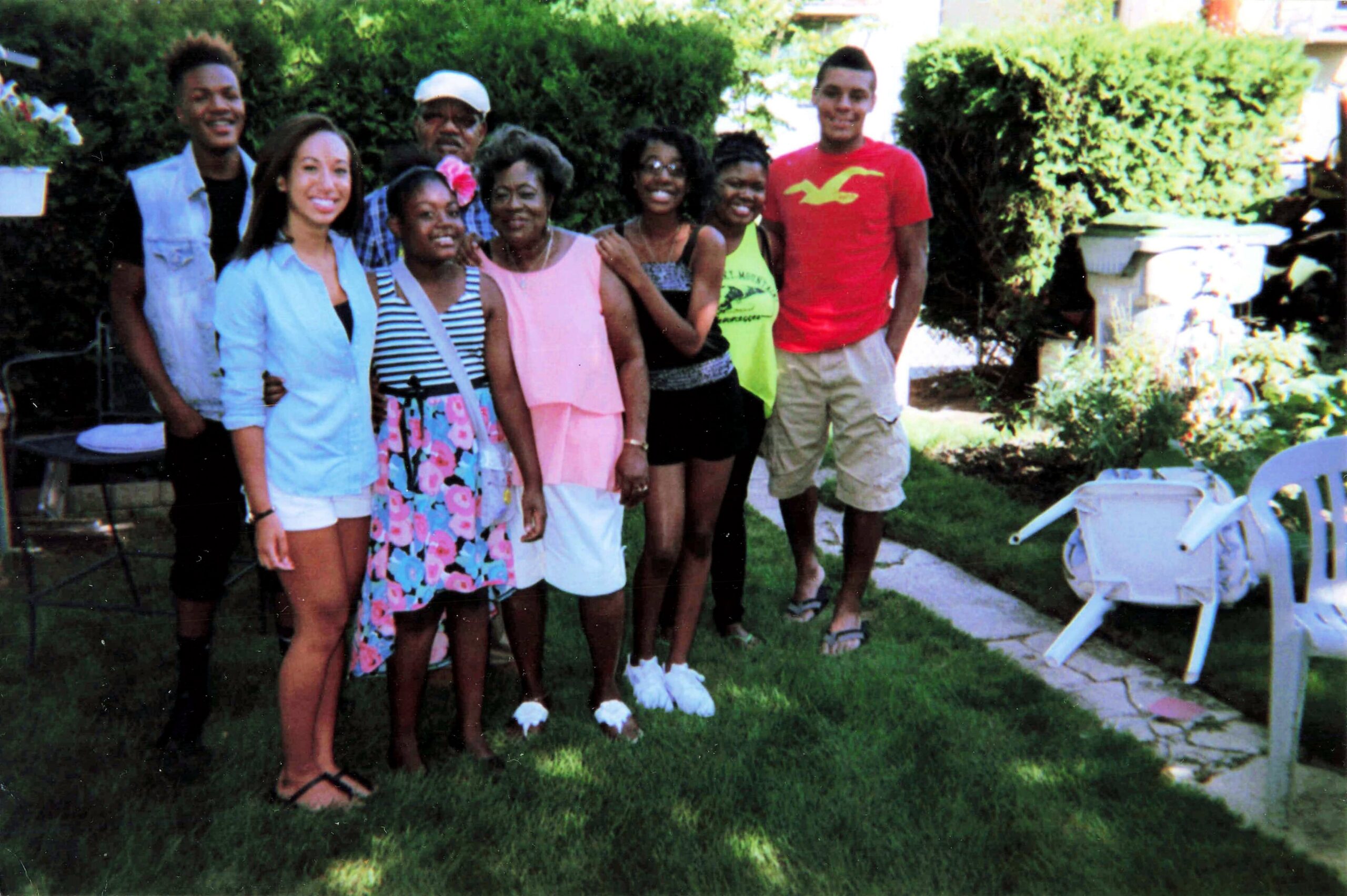 Group of eight people posing together in a sunny backyard, smiling, with hedges and lawn behind them.