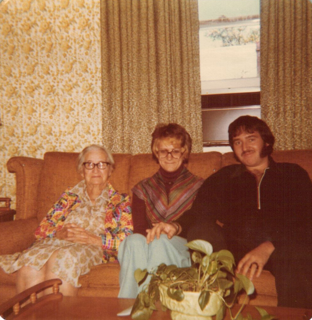 Three people sit on a brown couch in a vintage living room, elderly woman on left, middle-aged woman center, man on right.