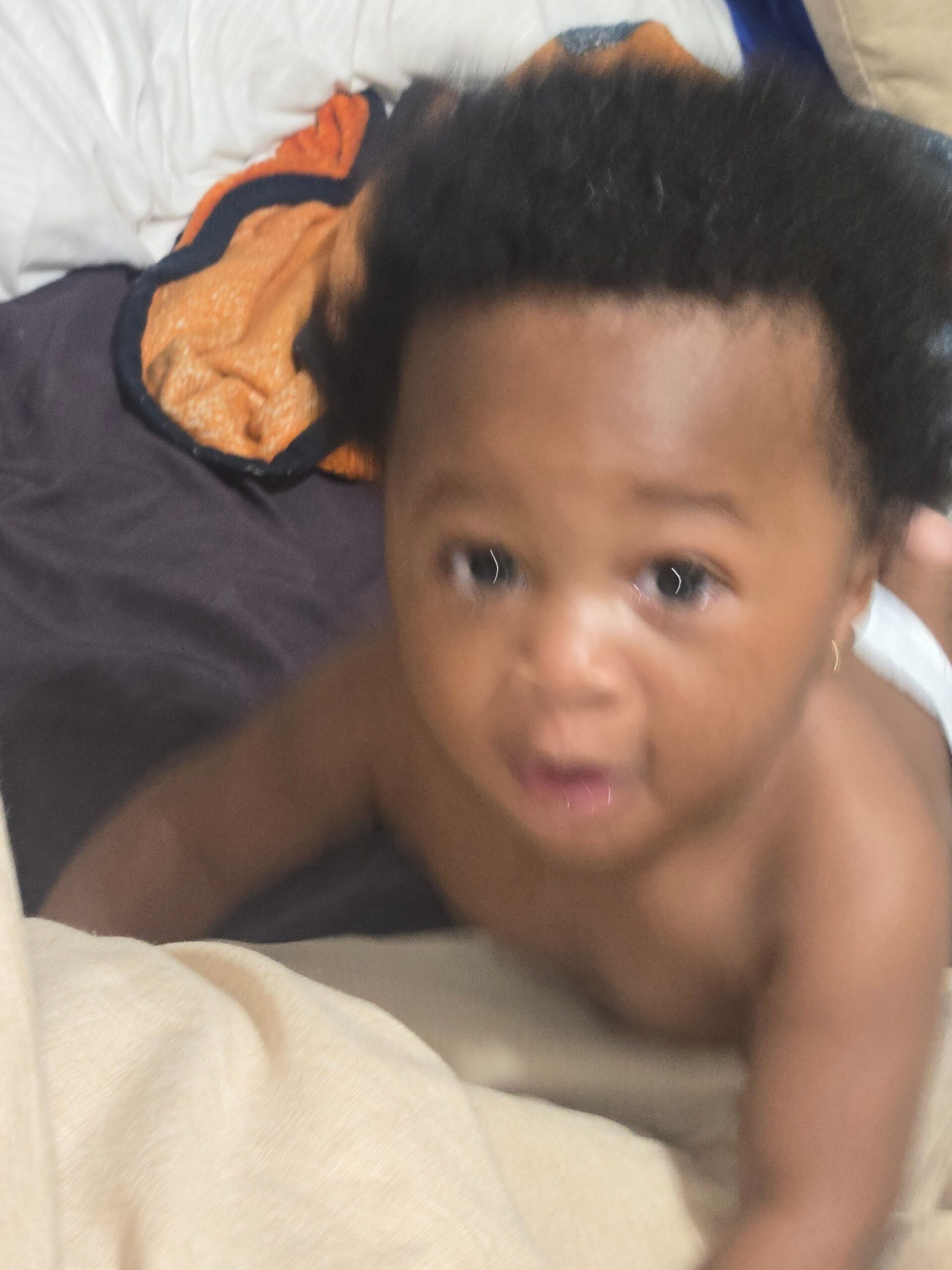 Close-up of a young child lying on a bed, looking toward the camera with wide eyes, an orange towel behind the head.