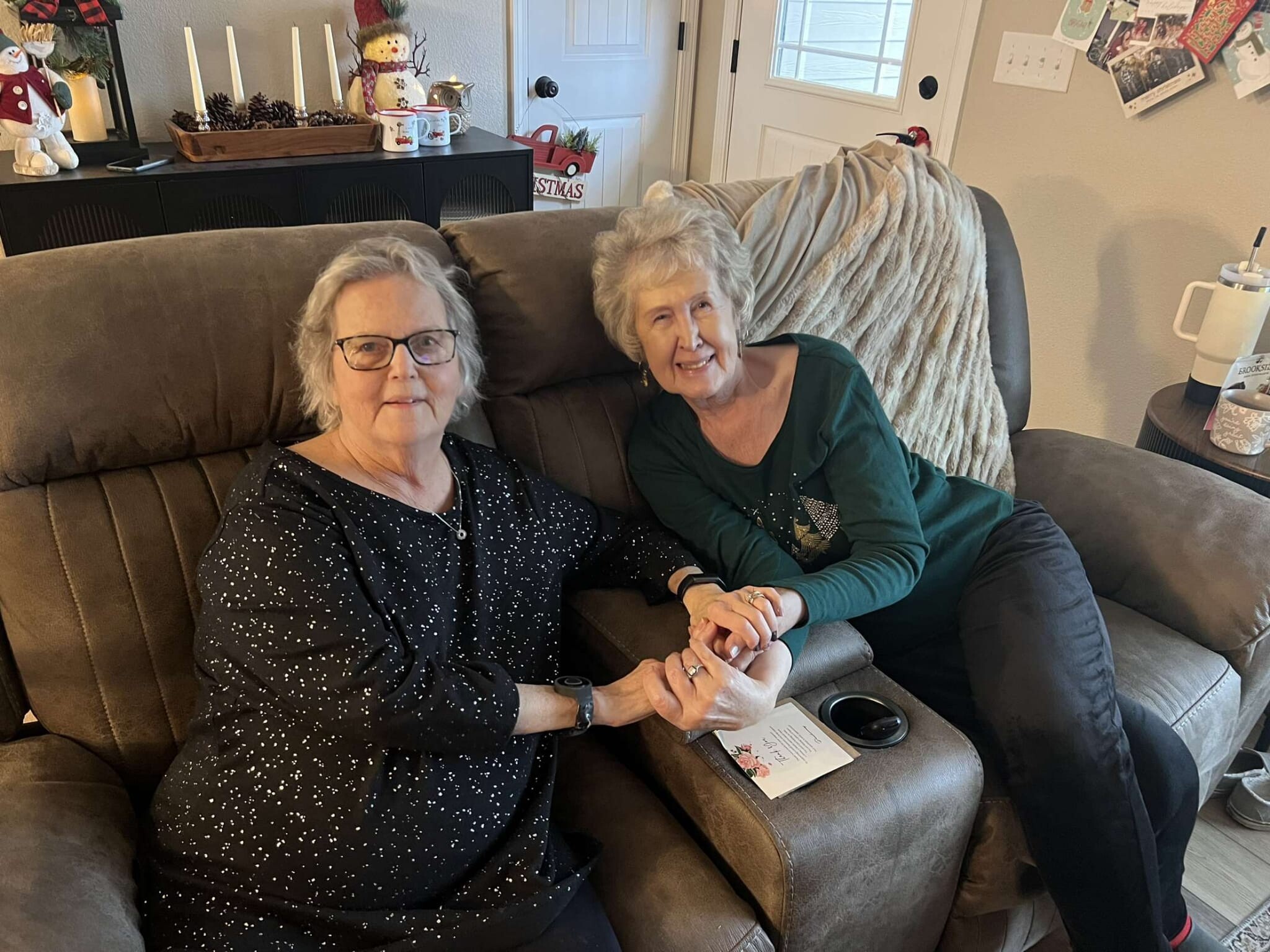 Two elderly women sit close together on a brown couch, holding hands and smiling, with Christmas decorations in the background.