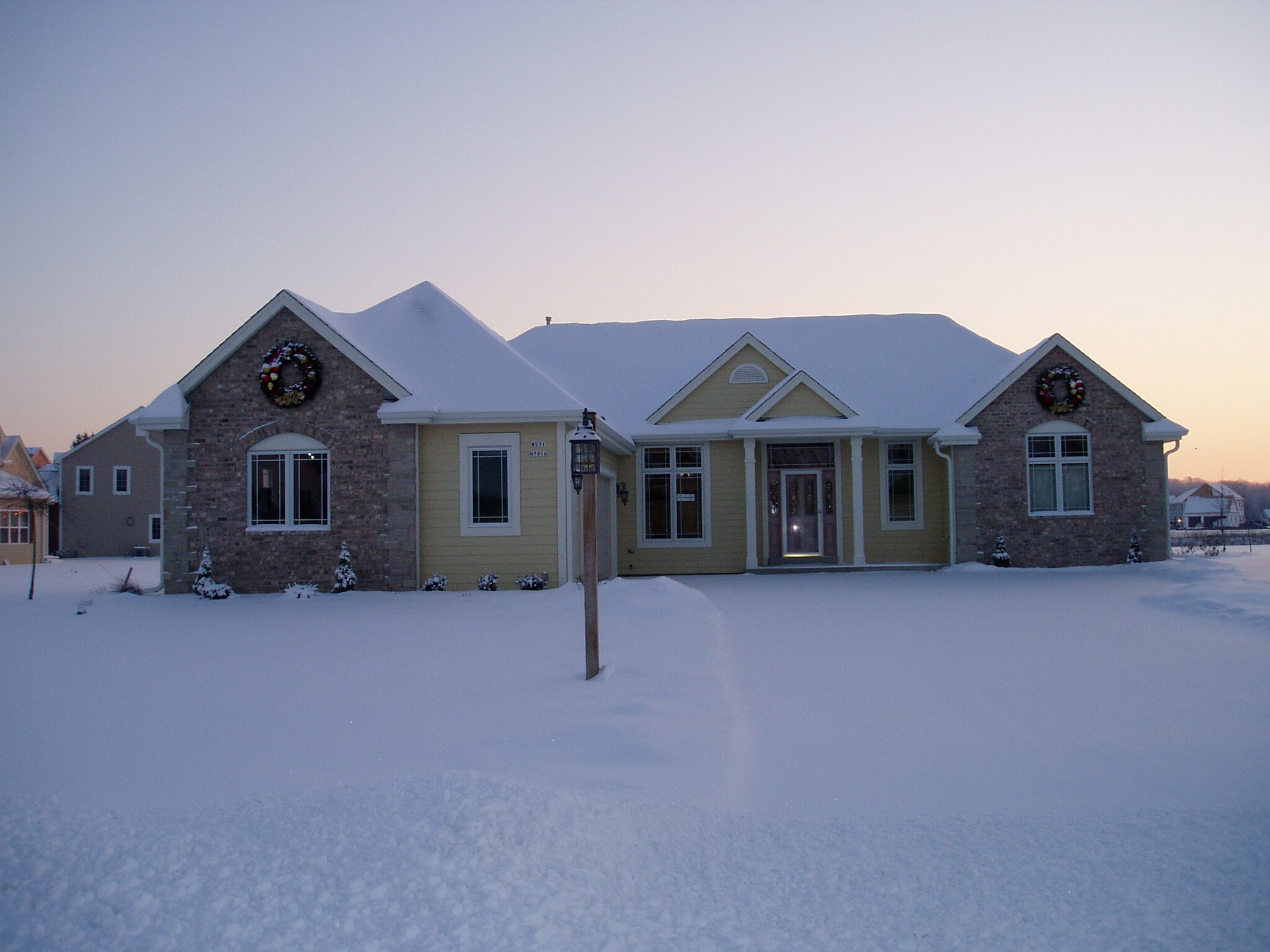 Snow-covered suburban street with two-will houses and wreaths on the façades at dusk.