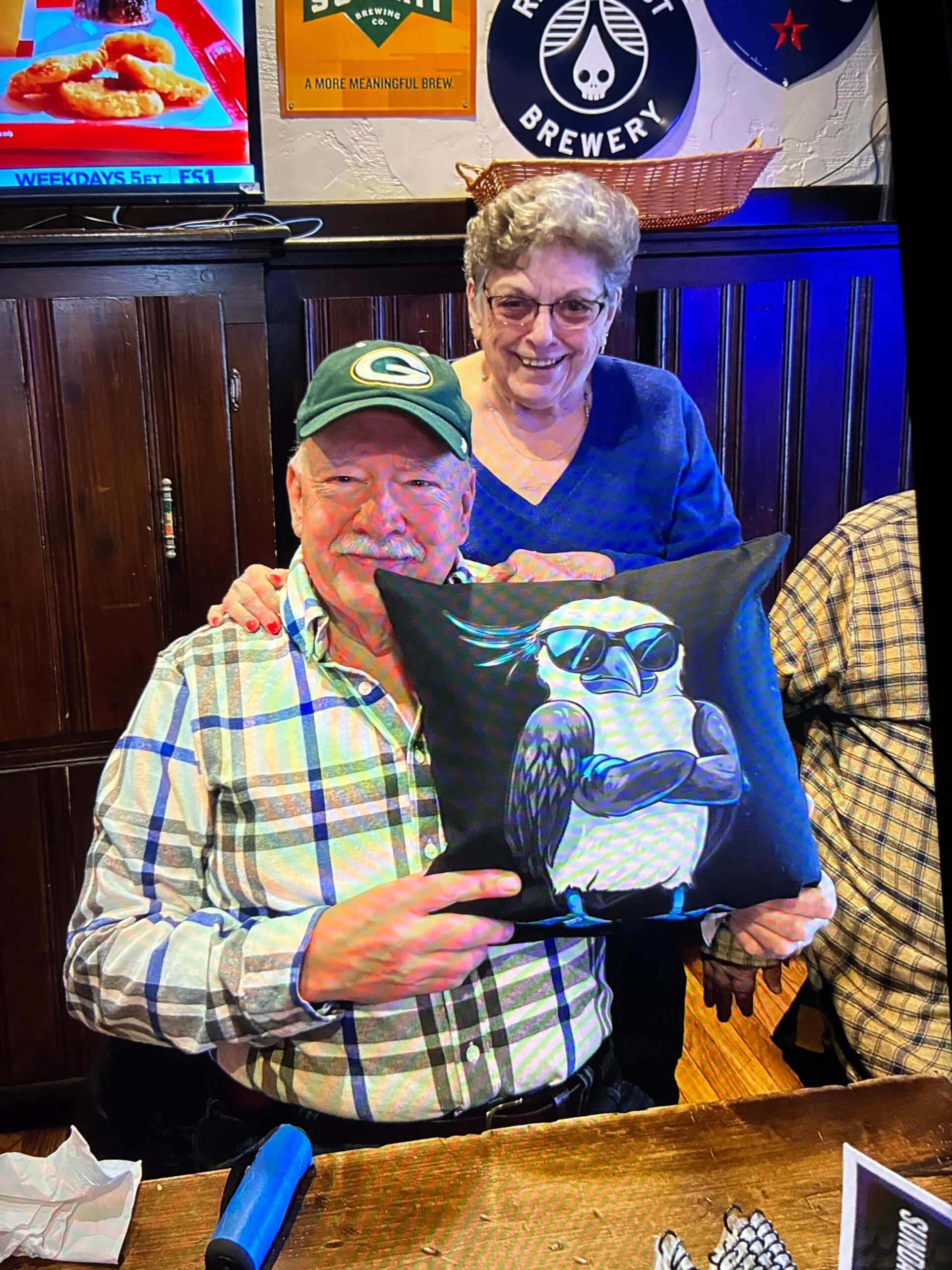 Smiling elderly couple in a pub: man in a plaid shirt and Green Bay Packers cap holds a penguin-printed pillow, as a woman in a blue sweater stands behind them.