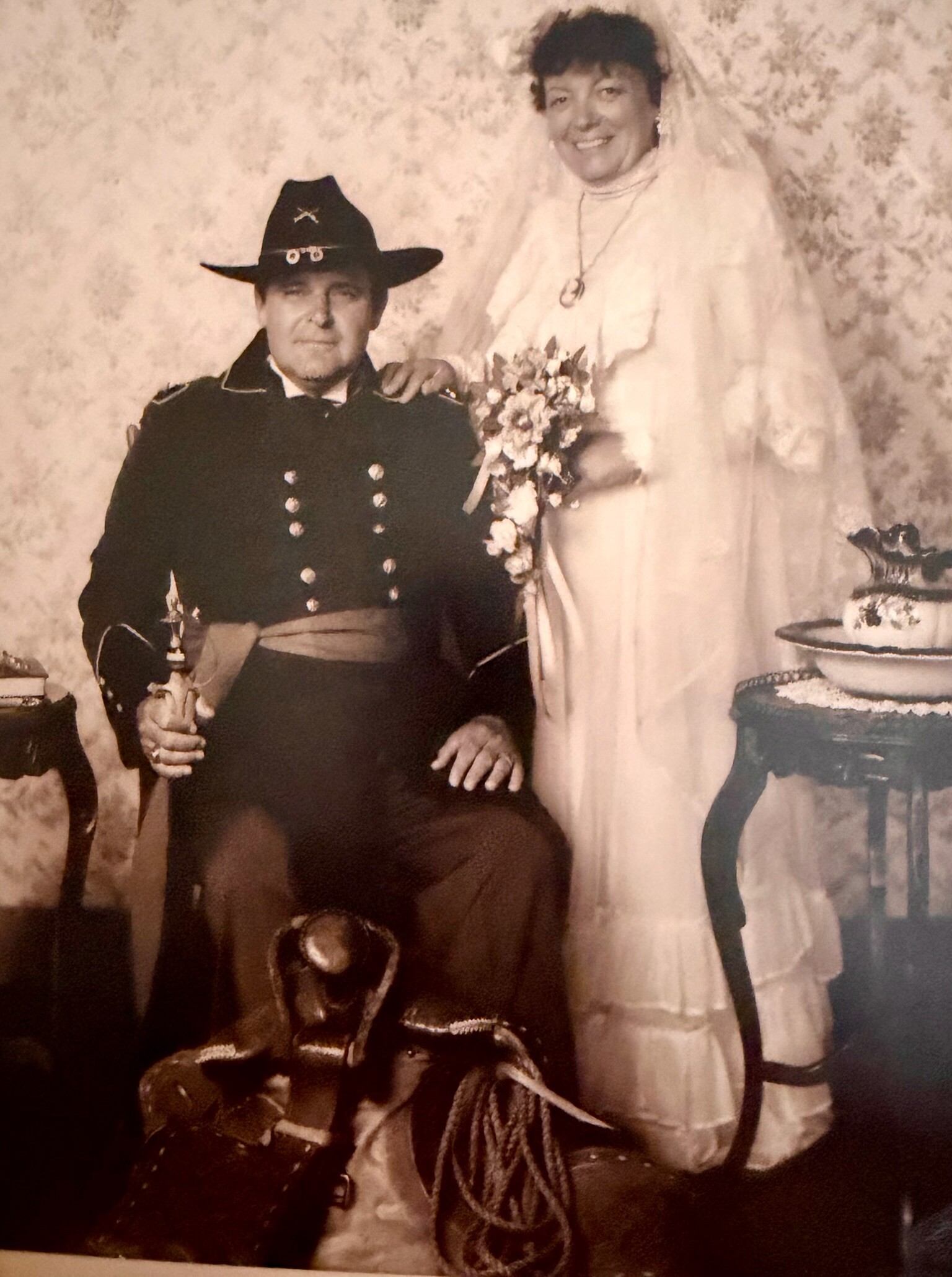 Bride in a white wedding dress standing next to a seated man in a dark double-breasted uniform and cowboy hat, holding a bouquet and posing for a vintage wedding portrait.