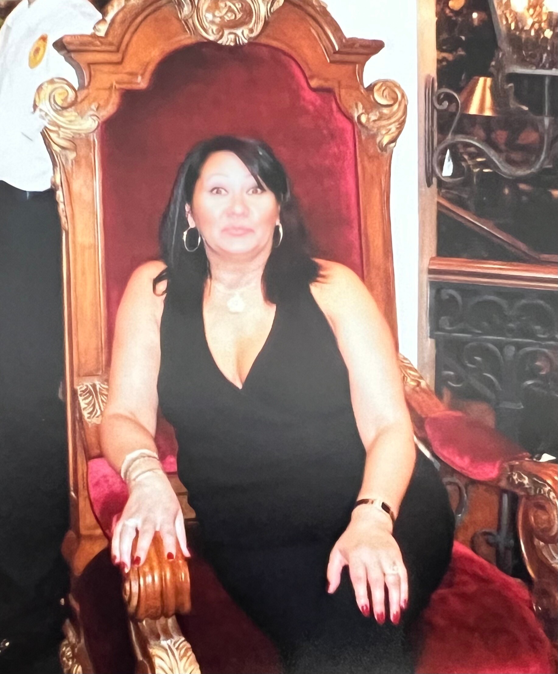 Woman in a black dress seated on an ornate gold throne with red velvet upholstery, hands resting on her lap.